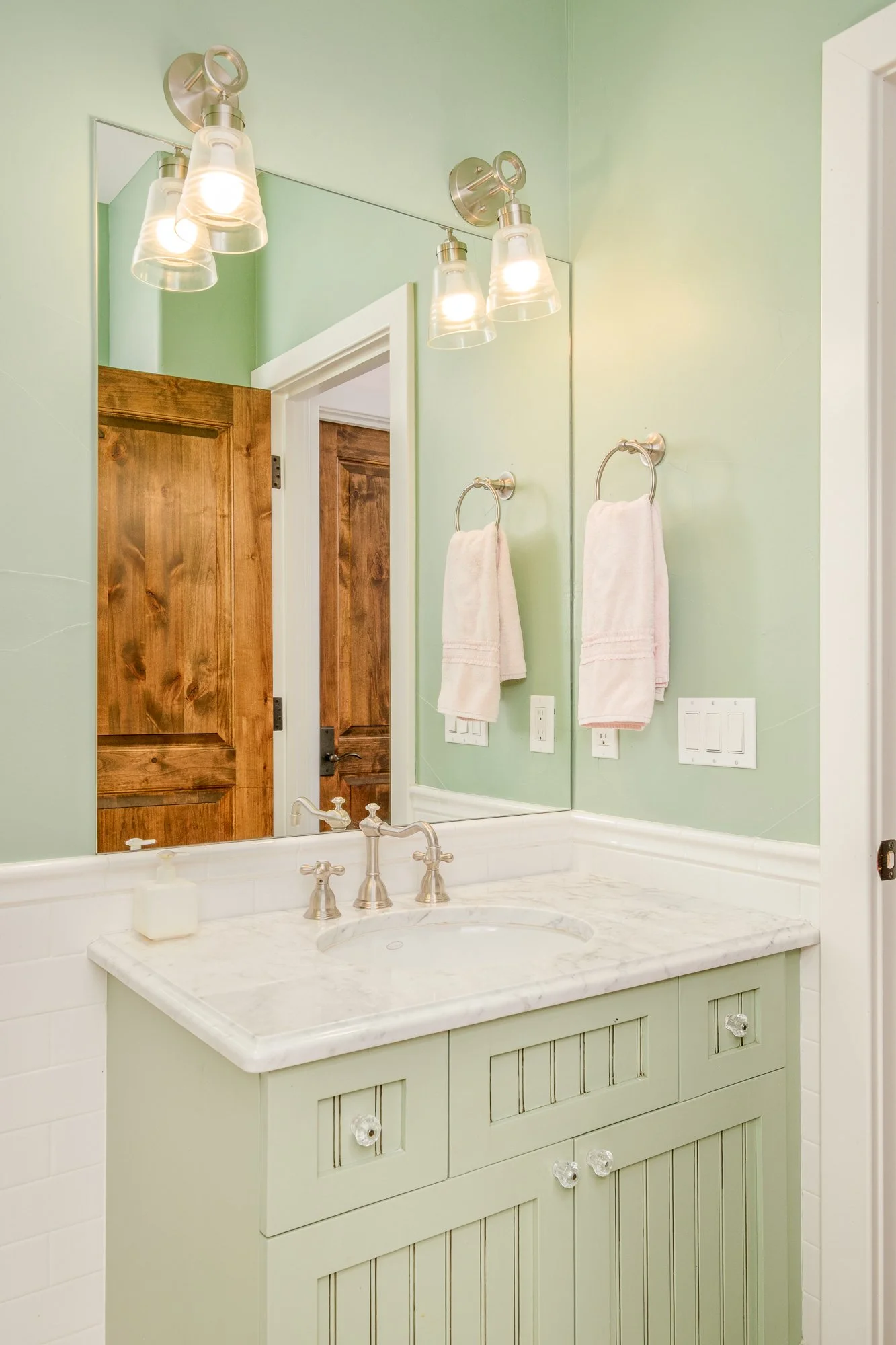 Mint green bathroom vanity with white marble countertop, framed mirror, brass faucet, towel rings, and illuminated glass sconces