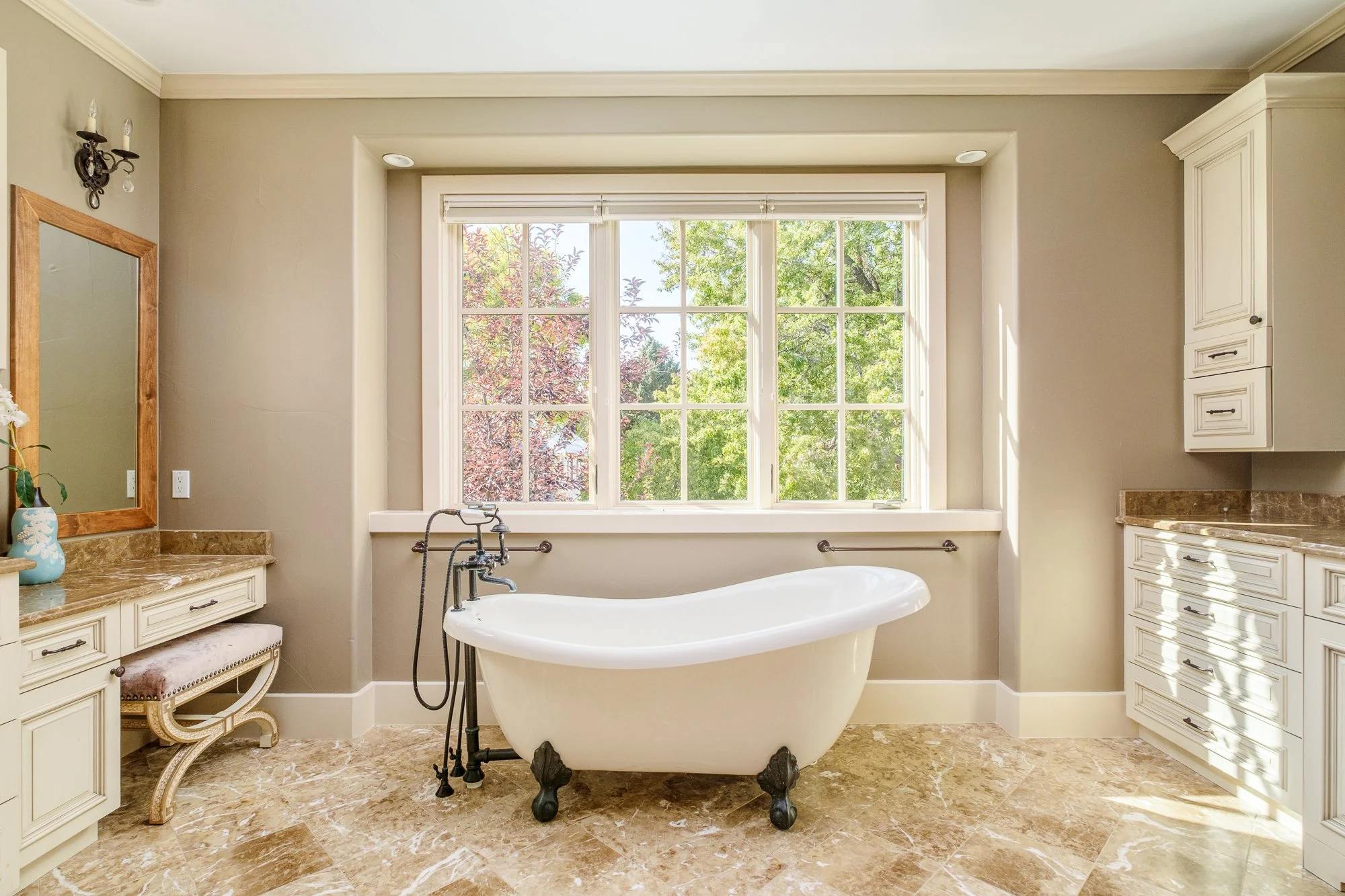 Elegant bathroom with freestanding clawfoot bathtub, large sunlit window, marble flooring, and white built-in cabinetry