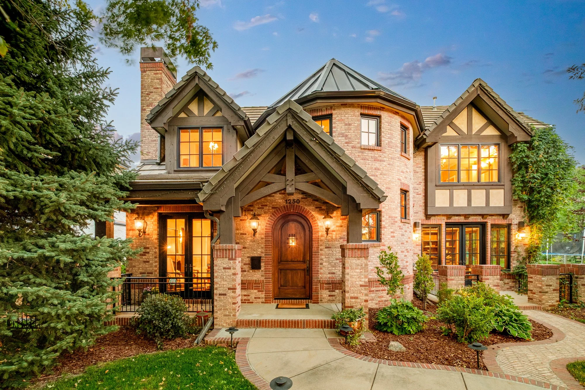 Large brick Tudor-style house with illuminated windows, landscaped front yard, and covered entrance at dusk