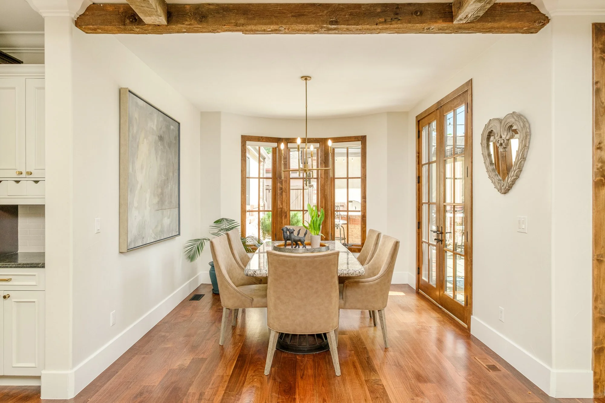 Bright dining room with a wood dining table, six upholstered chairs, modern chandelier, French doors, large windows, and exposed wood ceiling beams