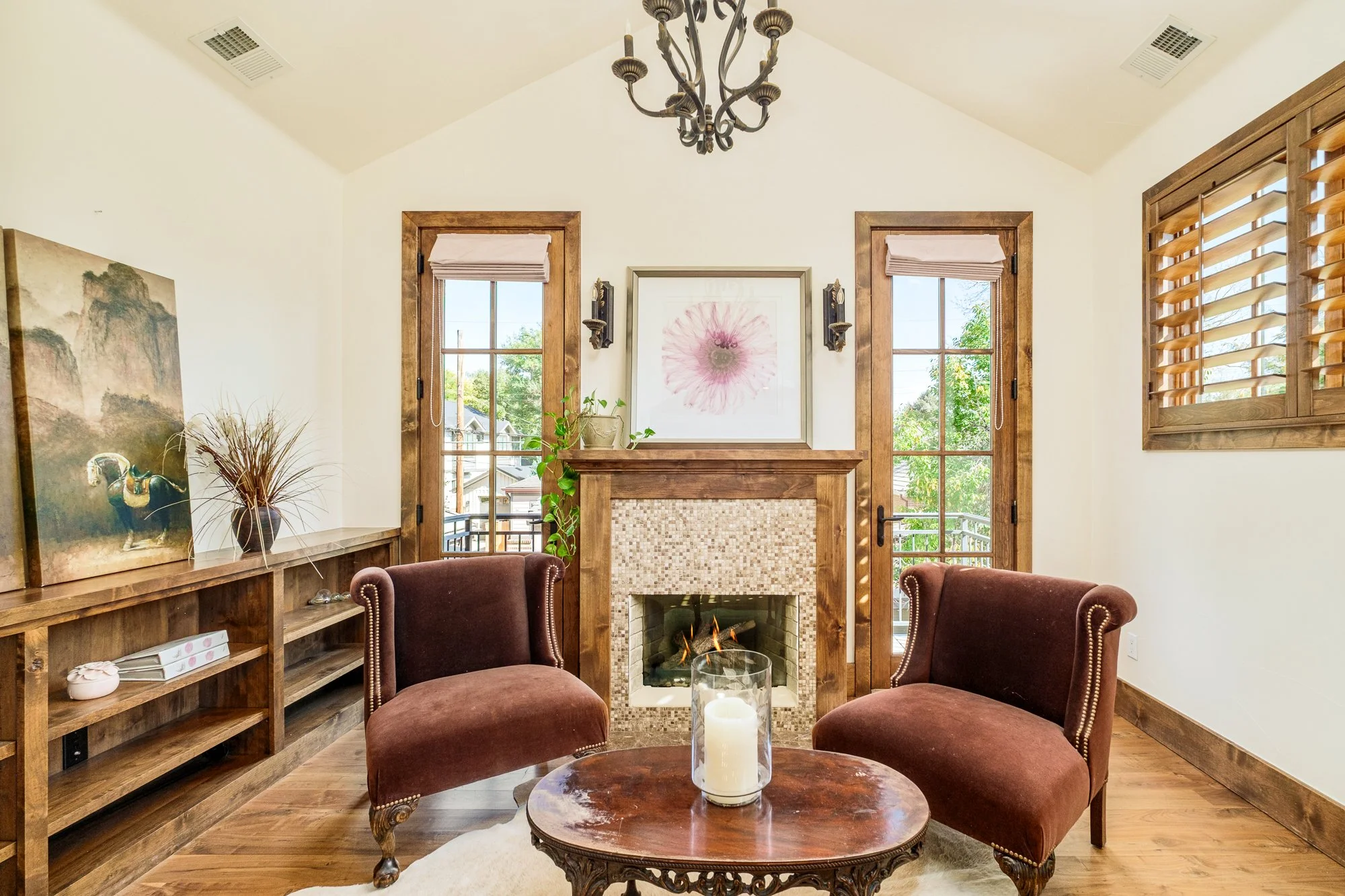 Elegant living room with a tiled fireplace, two burgundy armchairs, carved wood shelving, large windows, and a ceiling chandelier