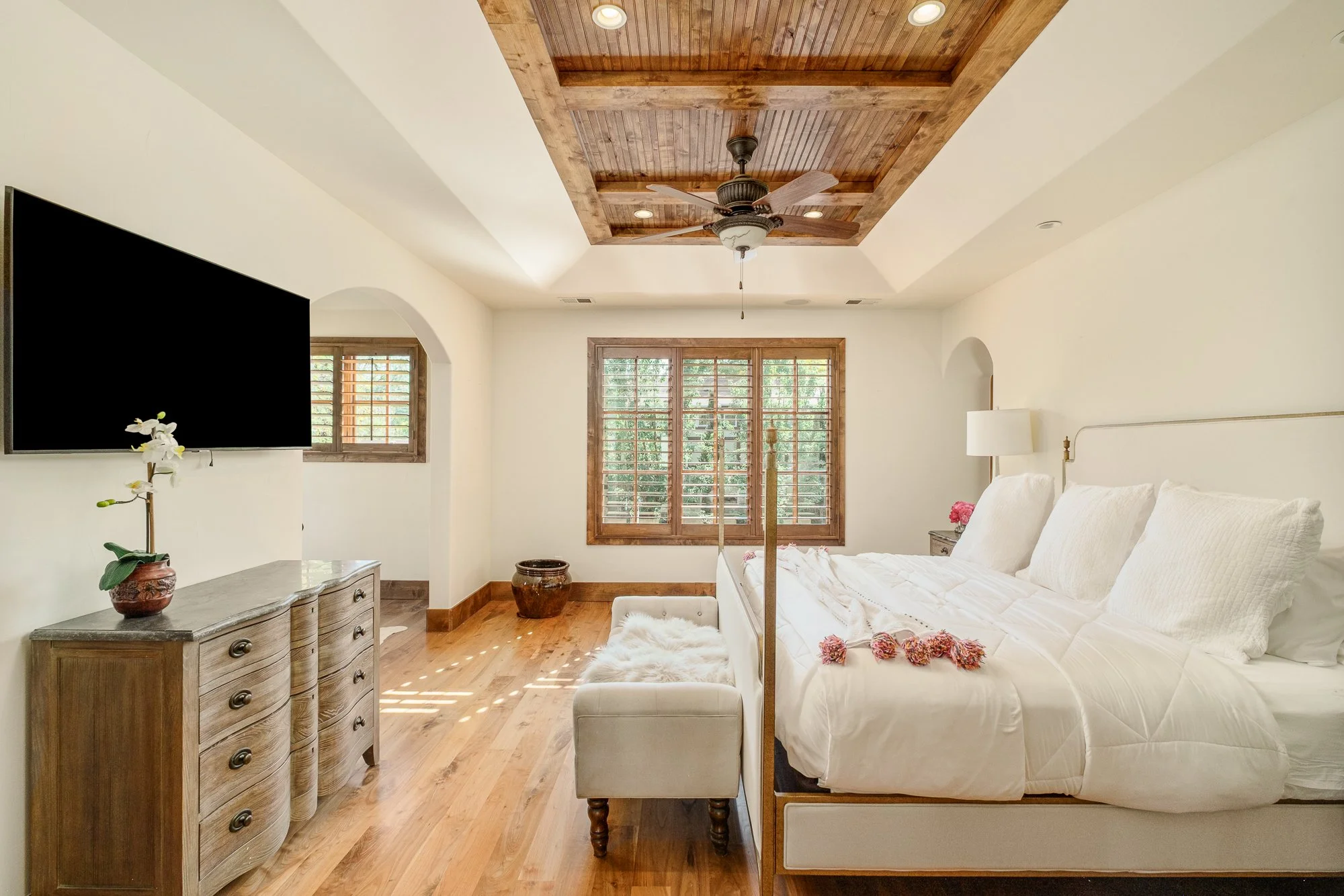 Bright bedroom with a four-poster bed, wood dresser, wall-mounted TV, plantation shutters, and rustic wood coffered ceiling