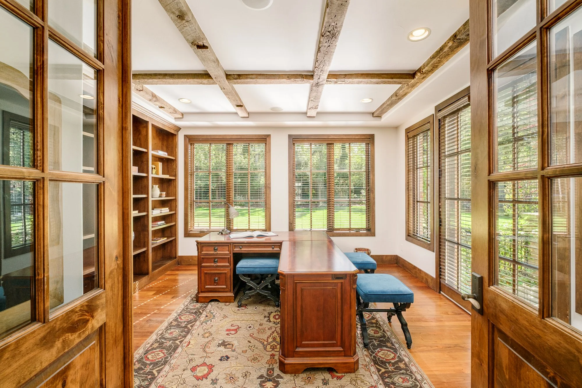Home office with a large wooden desk, built-in bookshelves, exposed beams, and windows overlooking a landscaped yard