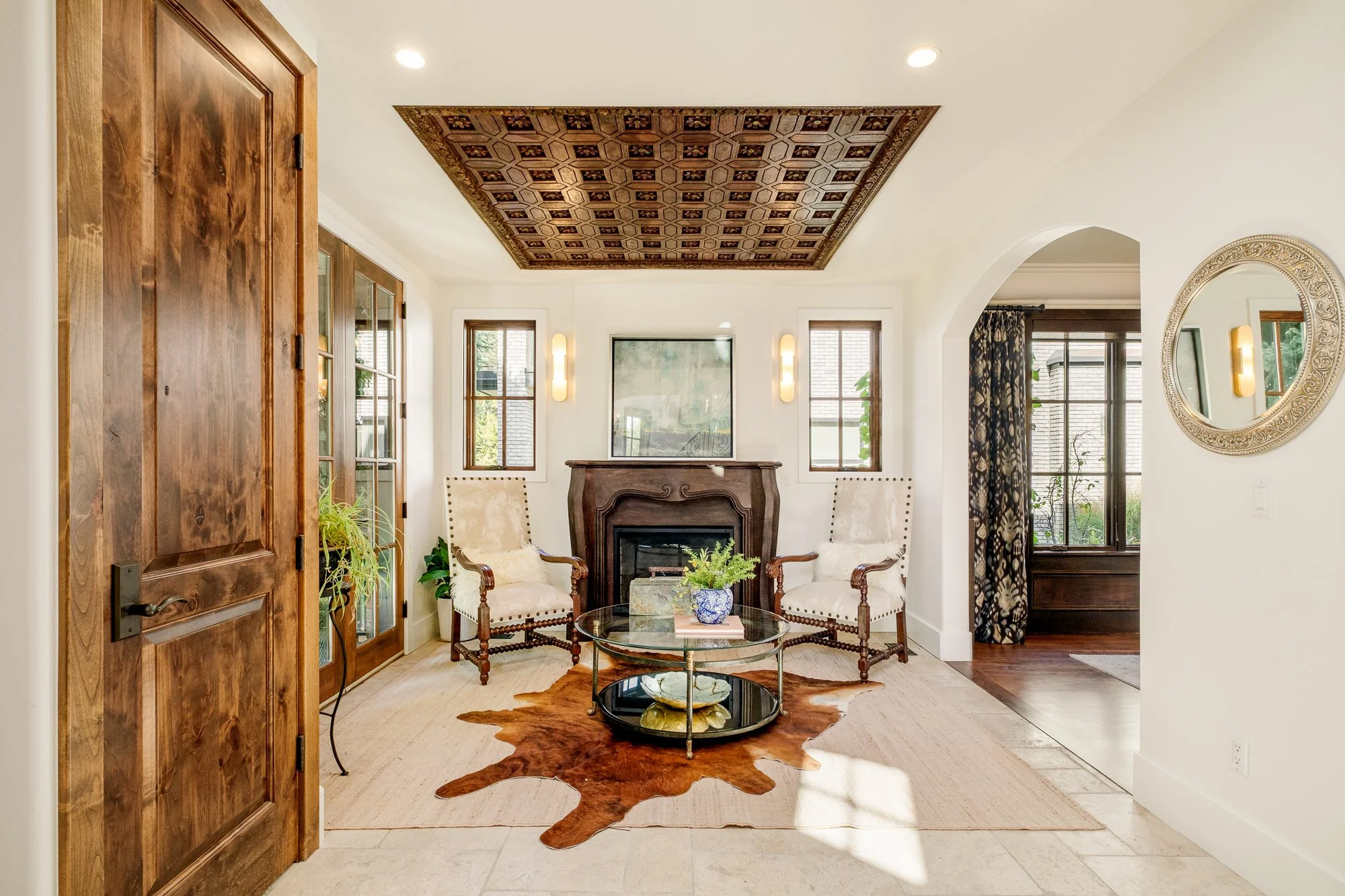 Elegant living room with a carved wood fireplace, two upholstered armchairs, glass coffee table, cowhide rug, ornate coffered ceiling, and arched doorway