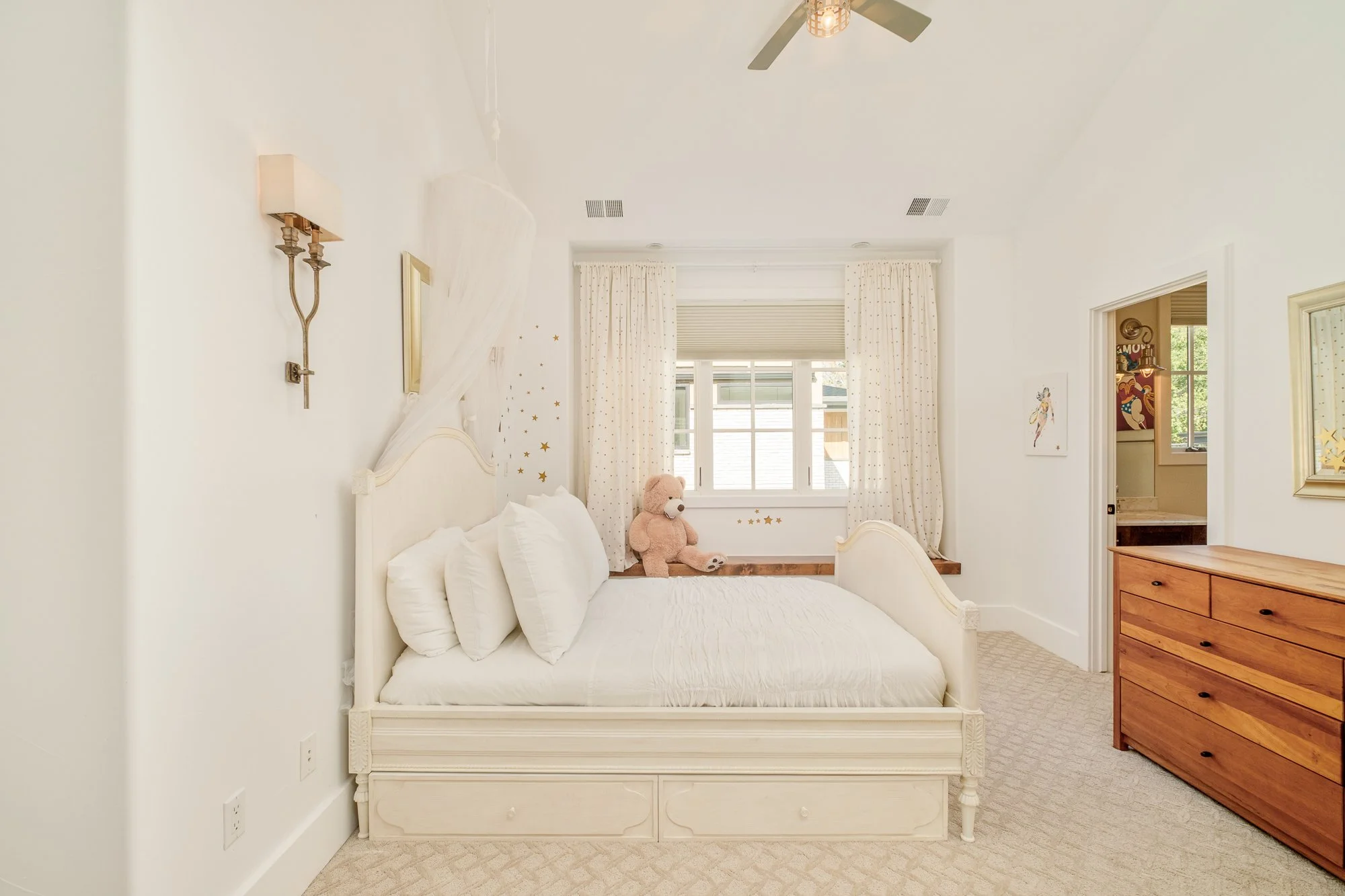 Bright white children’s bedroom with a vintage bed, teddy bear, star wall decals, window seat, and wooden dresser.