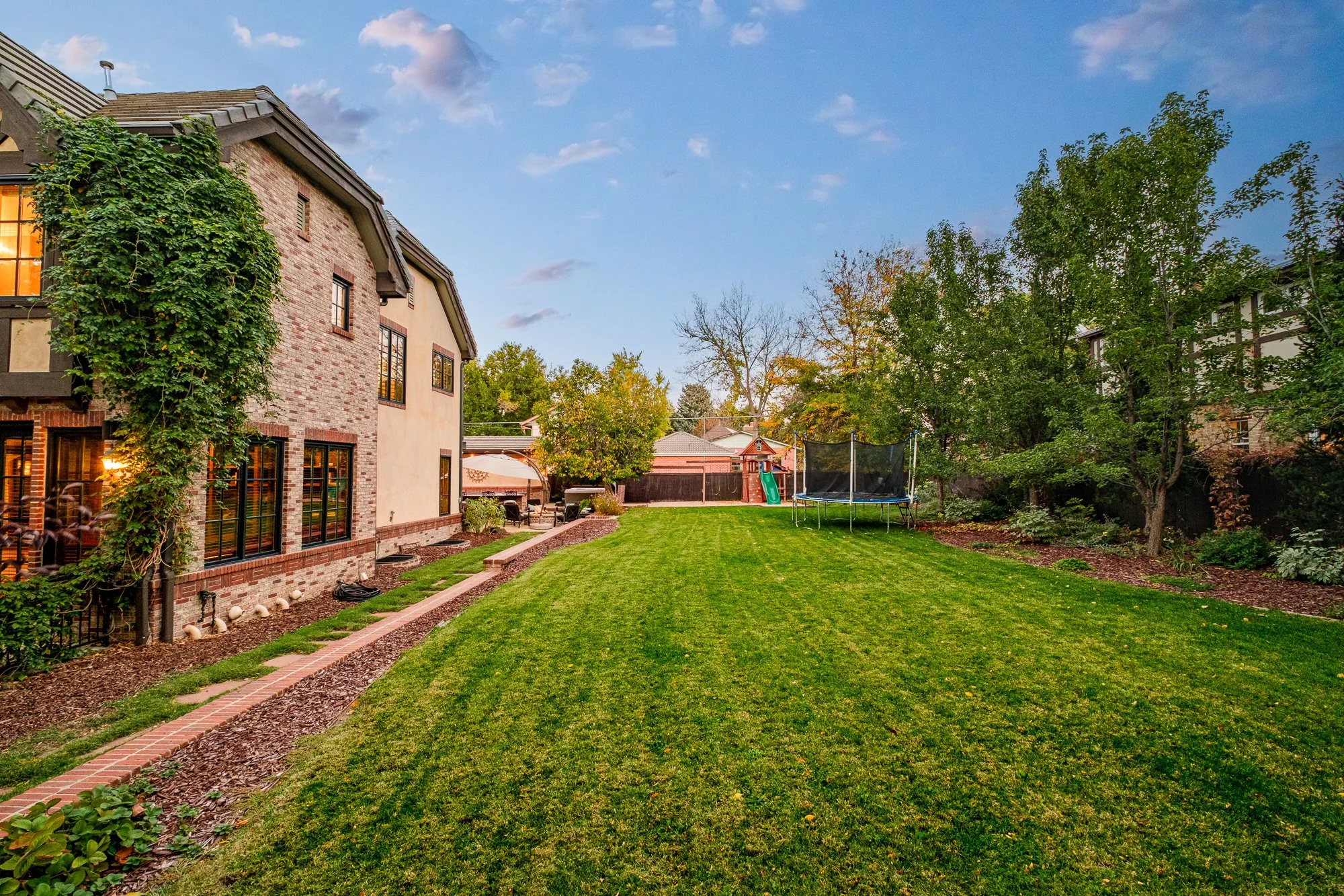 Spacious backyard with a brick house, manicured lawn, trampoline, playset, patio, and mature trees