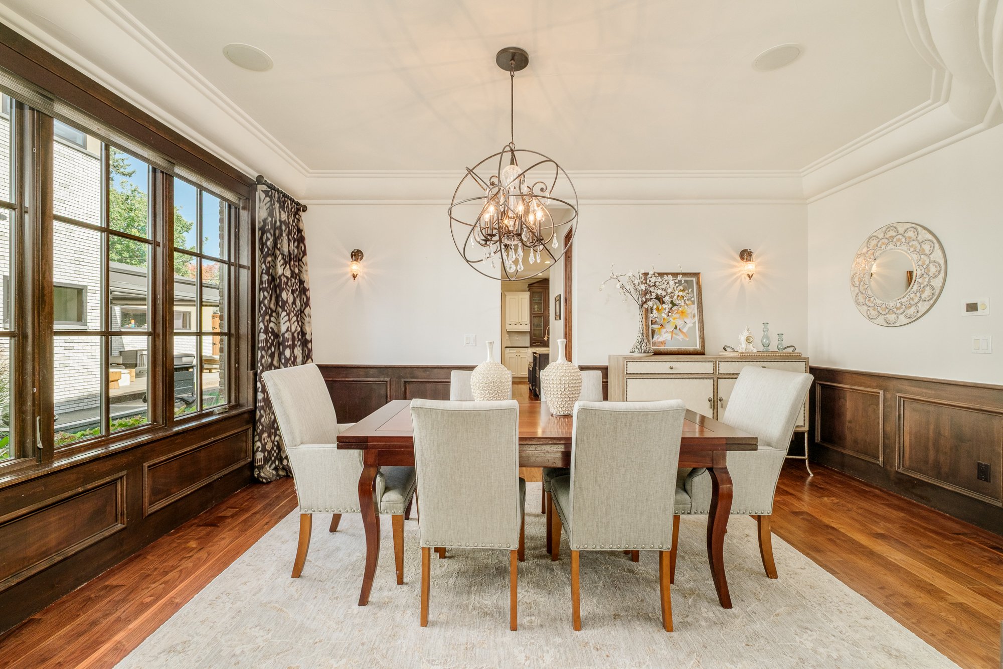 Elegant dining room with a wood table, six upholstered chairs, ornate chandelier, large windows, and dark wood paneling