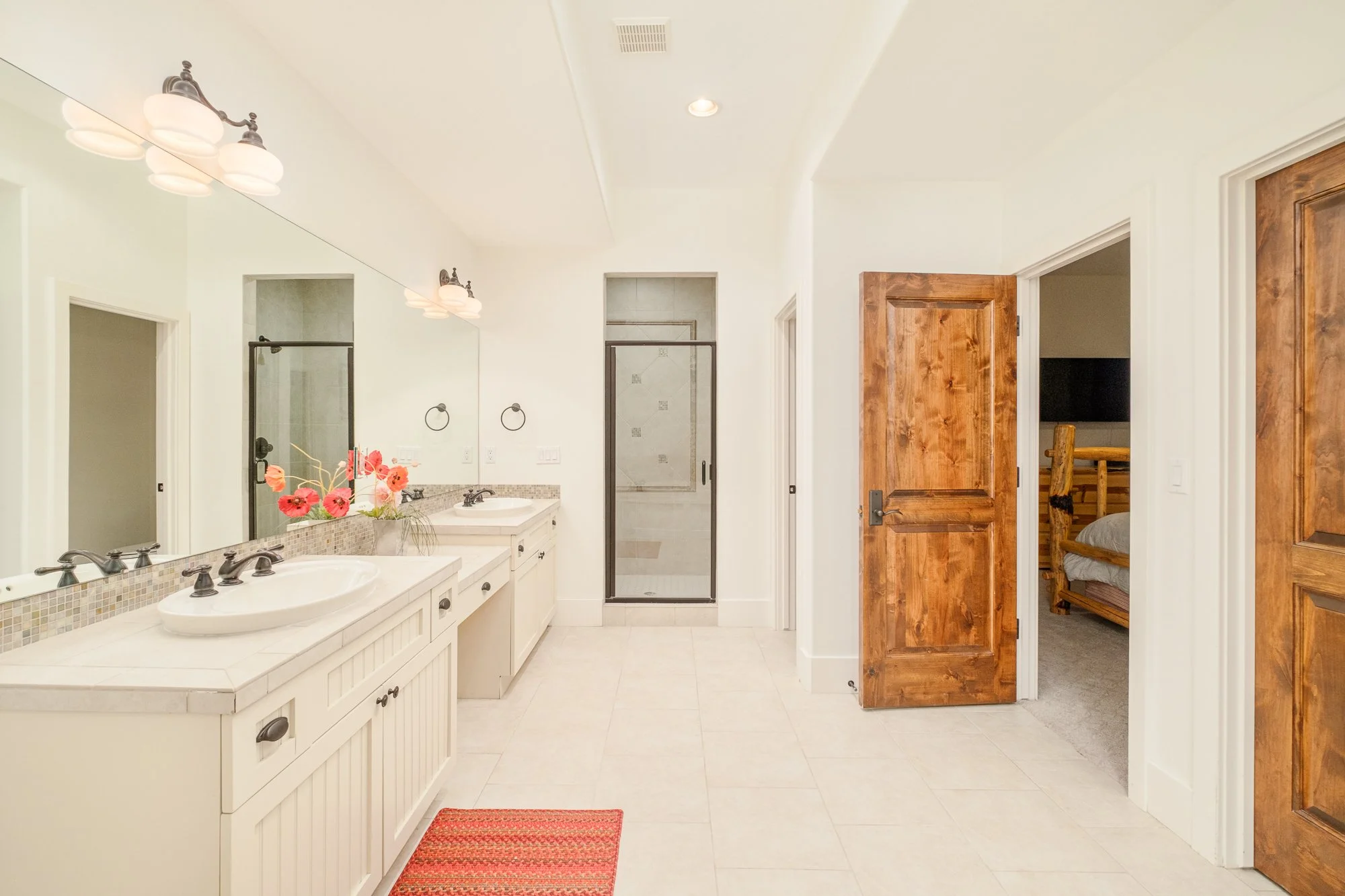 Bright white bathroom with double vanity, large wall mirror, glass-enclosed shower, tiled floor, and rustic wooden doors