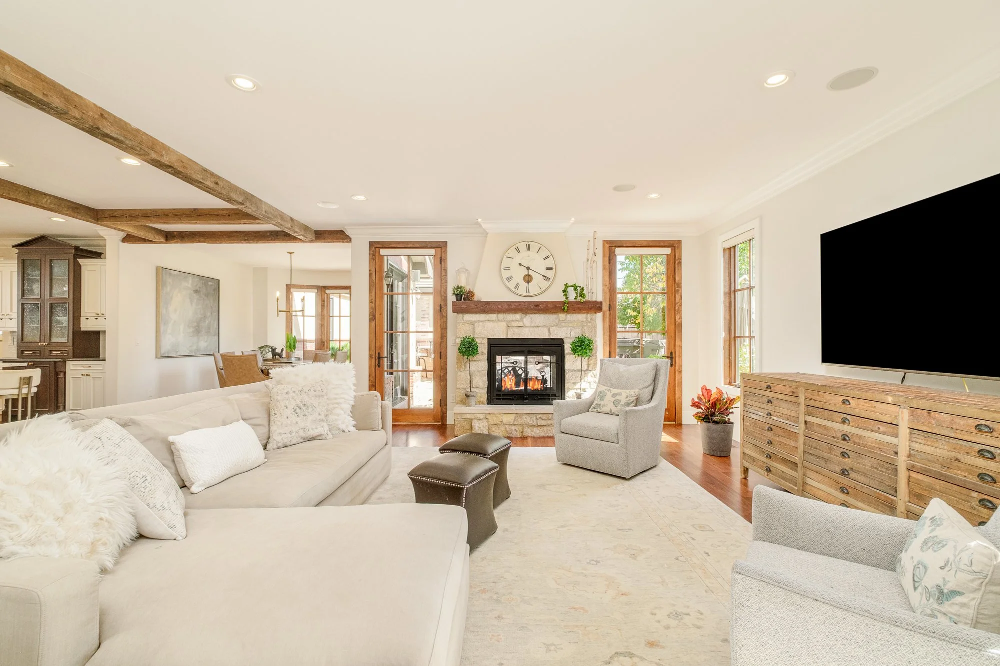 Bright open-concept living room with a stone fireplace, large wall-mounted TV, sectional sofa, armchairs, exposed wood beams, and adjacent kitchen.