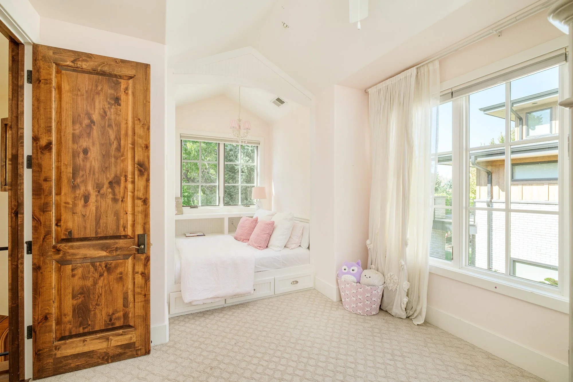 Bright children's bedroom with a built-in window seat, pink pillows, plush toys, hardwood door, and large windows.