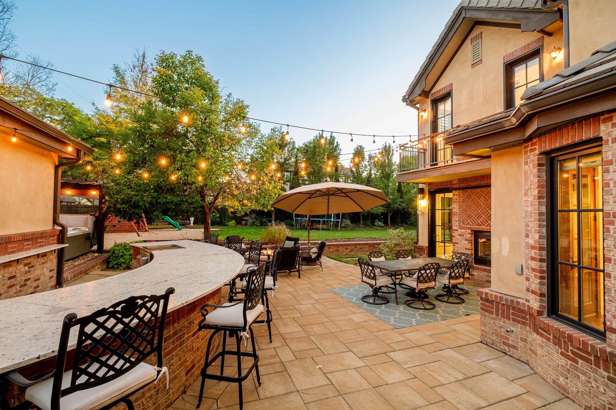 Spacious backyard patio with outdoor kitchen, bar seating, dining furniture, fire pit, umbrella, lawn, and string lights beside a brick-and-stucco home.