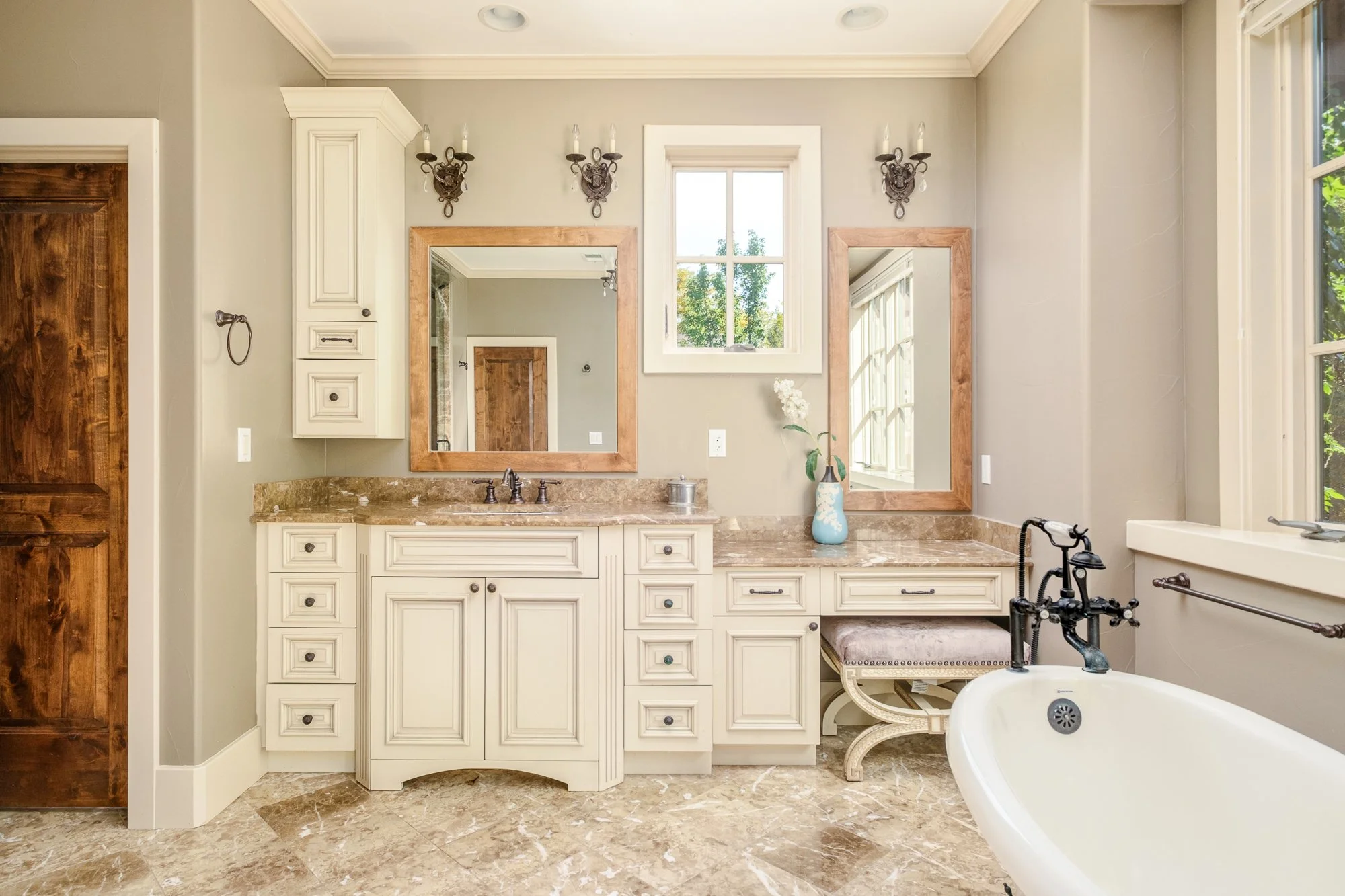 Elegant beige bathroom with cream vanity, marble countertops, framed mirrors, freestanding bathtub, and large windows