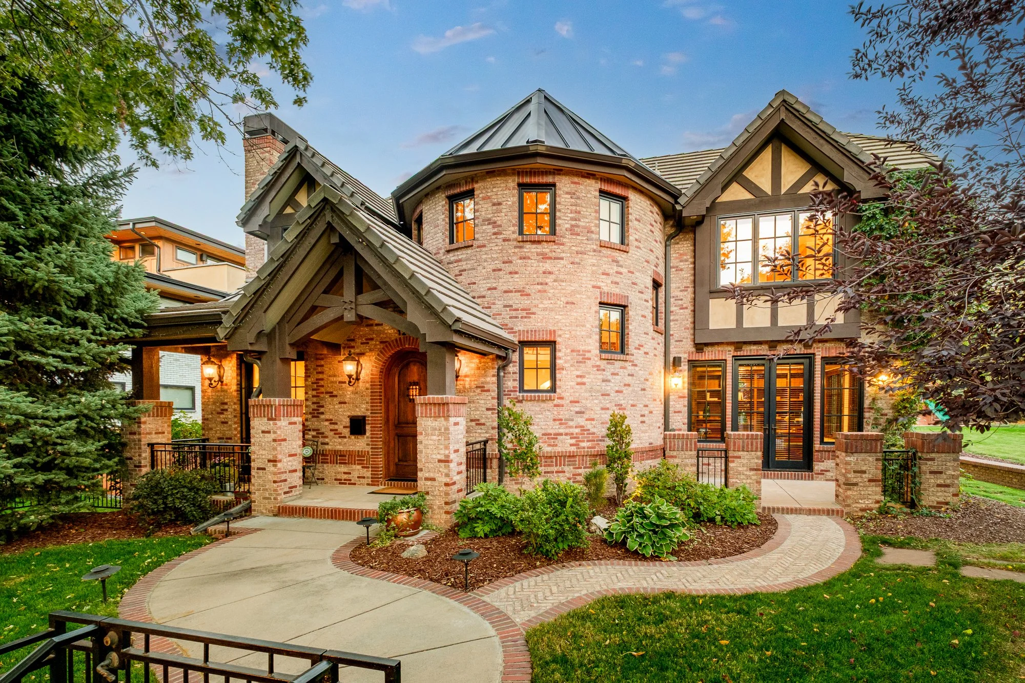 Large brick Tudor-style home with covered entry, turret roof, landscaped garden, and curved brick walkway