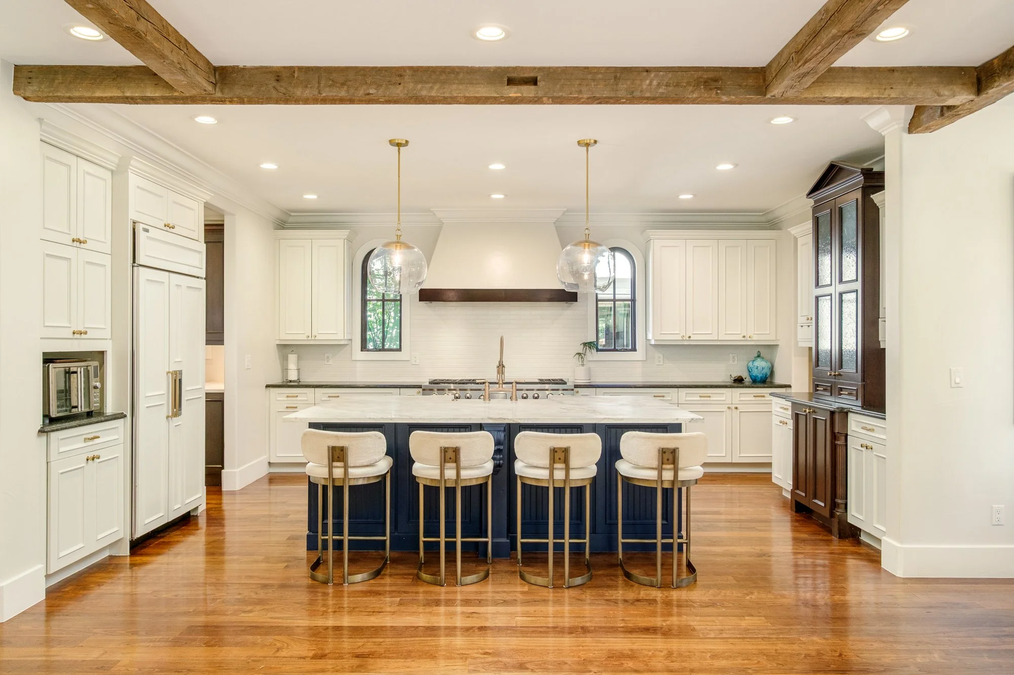 Spacious white kitchen with navy island, marble countertop, four upholstered bar stools, pendant lights, exposed wood beams, and stainless steel appliances.