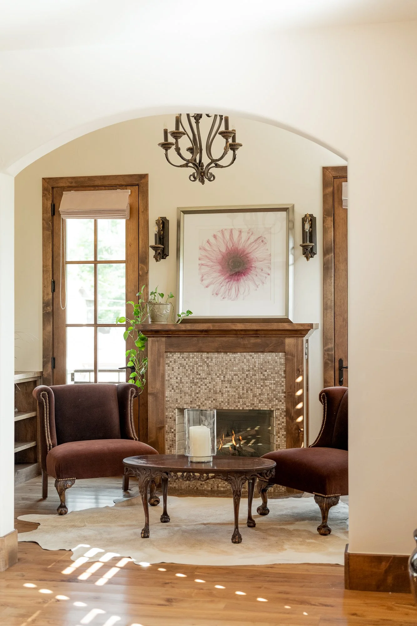 Elegant living room with a tiled fireplace, two burgundy wingback chairs, chandelier, framed floral artwork, and wood coffee table.