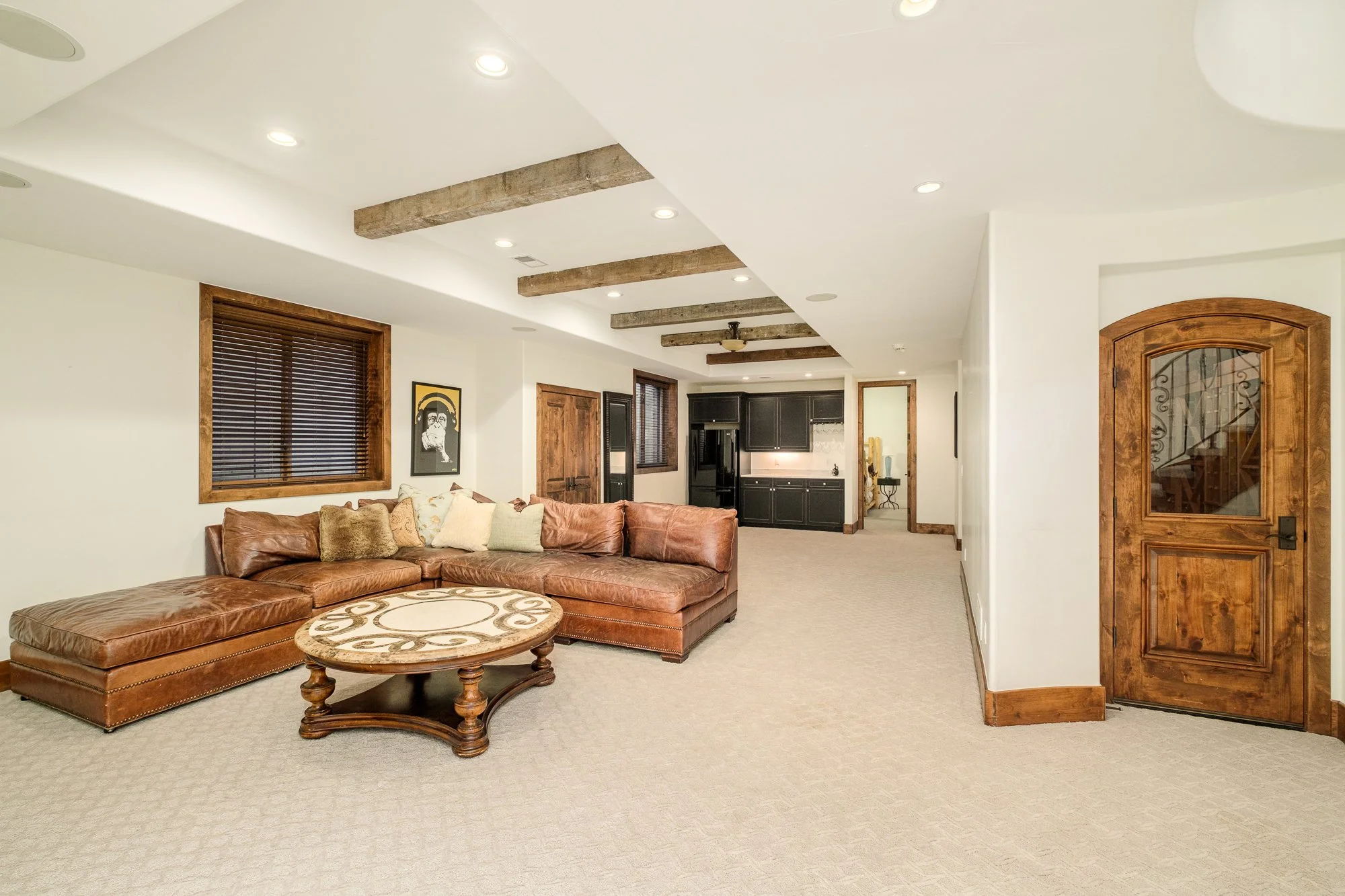 Spacious living room with a brown leather sectional, ornate coffee table, exposed wood ceiling beams, and adjacent kitchen.
