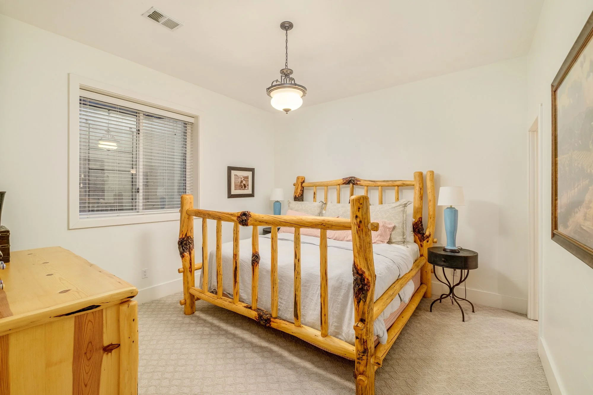 Rustic bedroom with a log-style wooden bed, white bedding, nightstands, blue lamps, window blinds, and light carpet