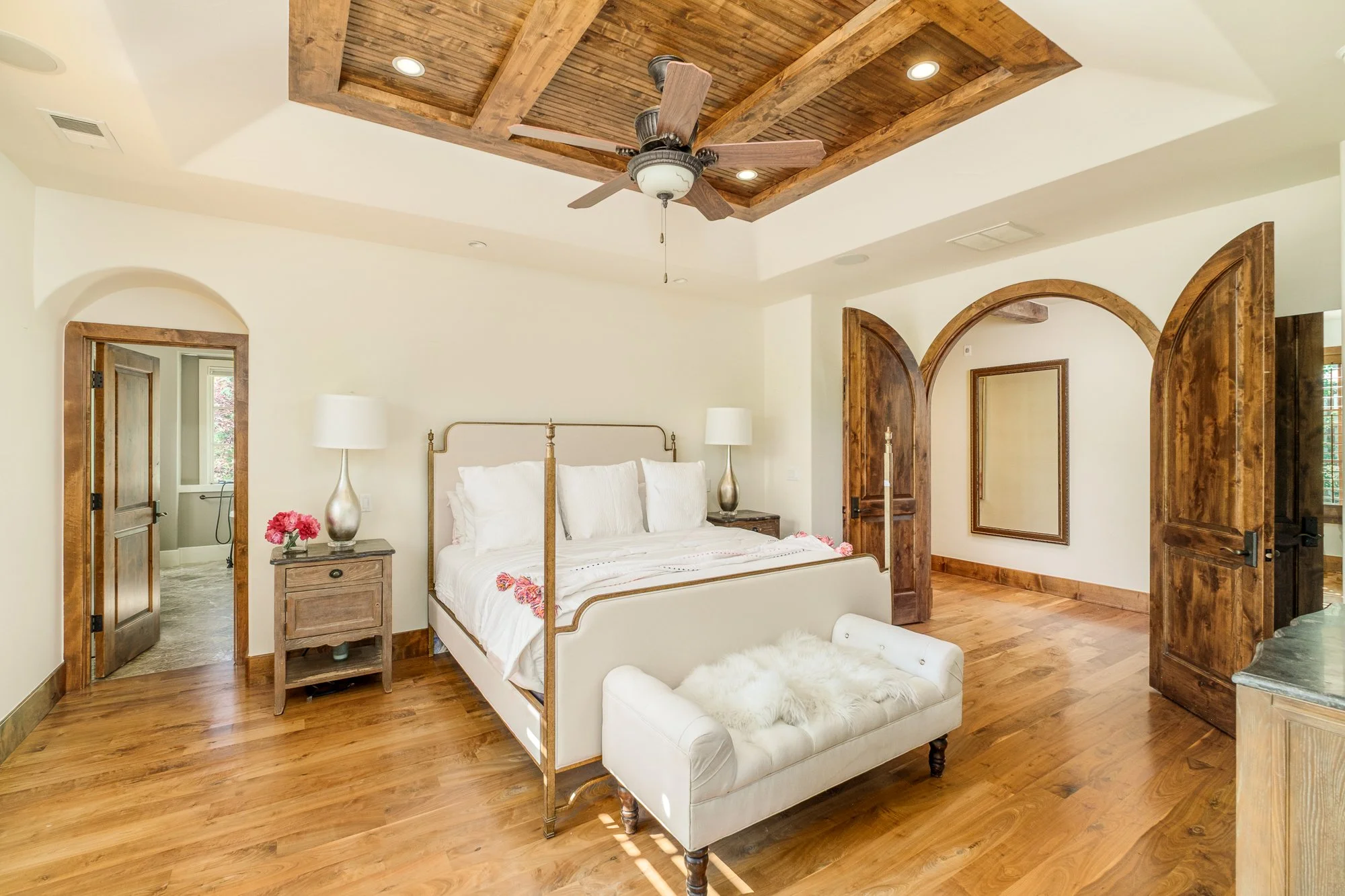 Elegant bedroom with a four-poster bed, white bedding, wood floors, arched doorways, and a rustic wood-beamed ceiling fan.