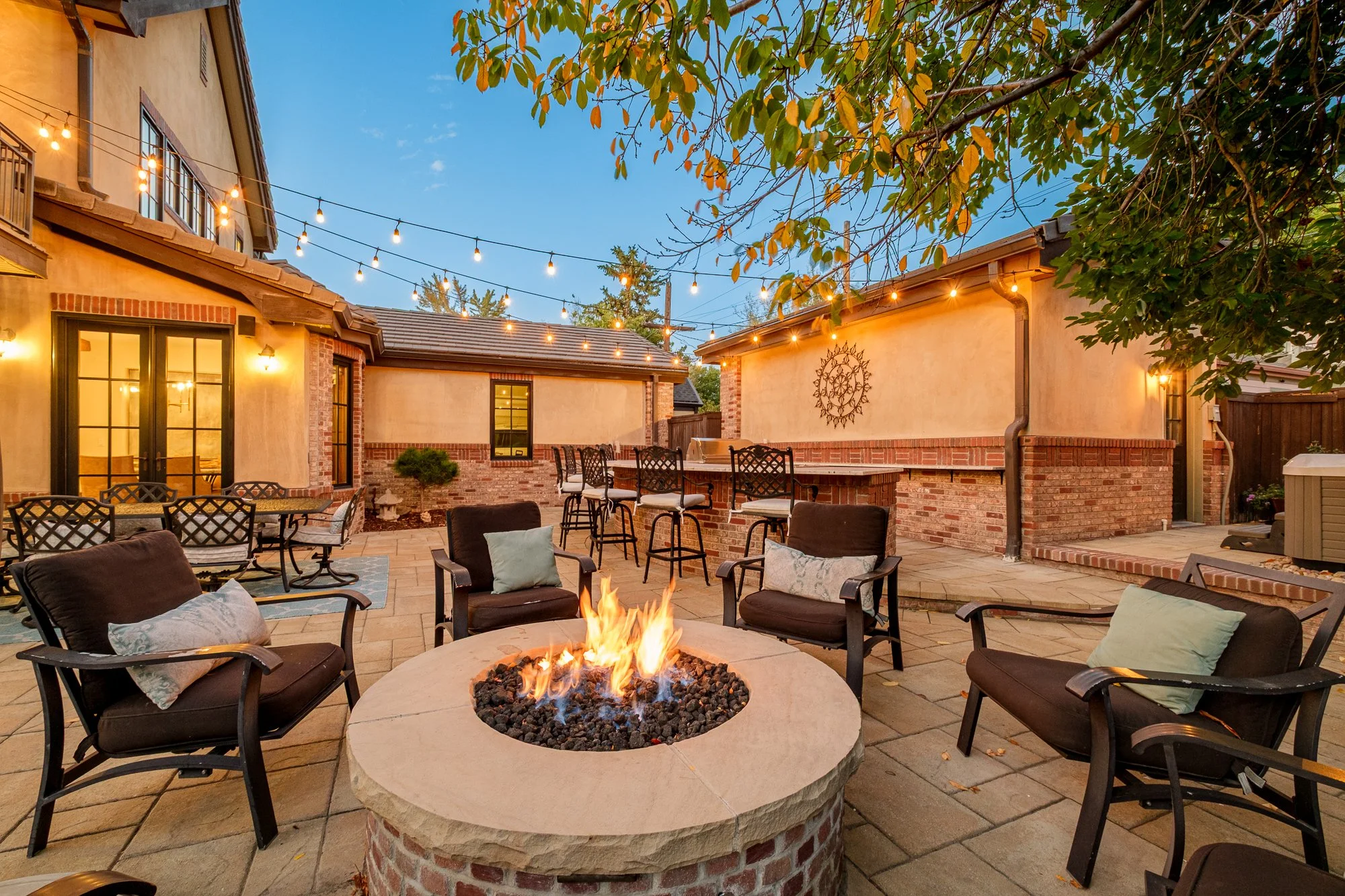 Cozy outdoor patio with a lit fire pit, cushioned chairs, bar seating, string lights, and stucco walls at dusk