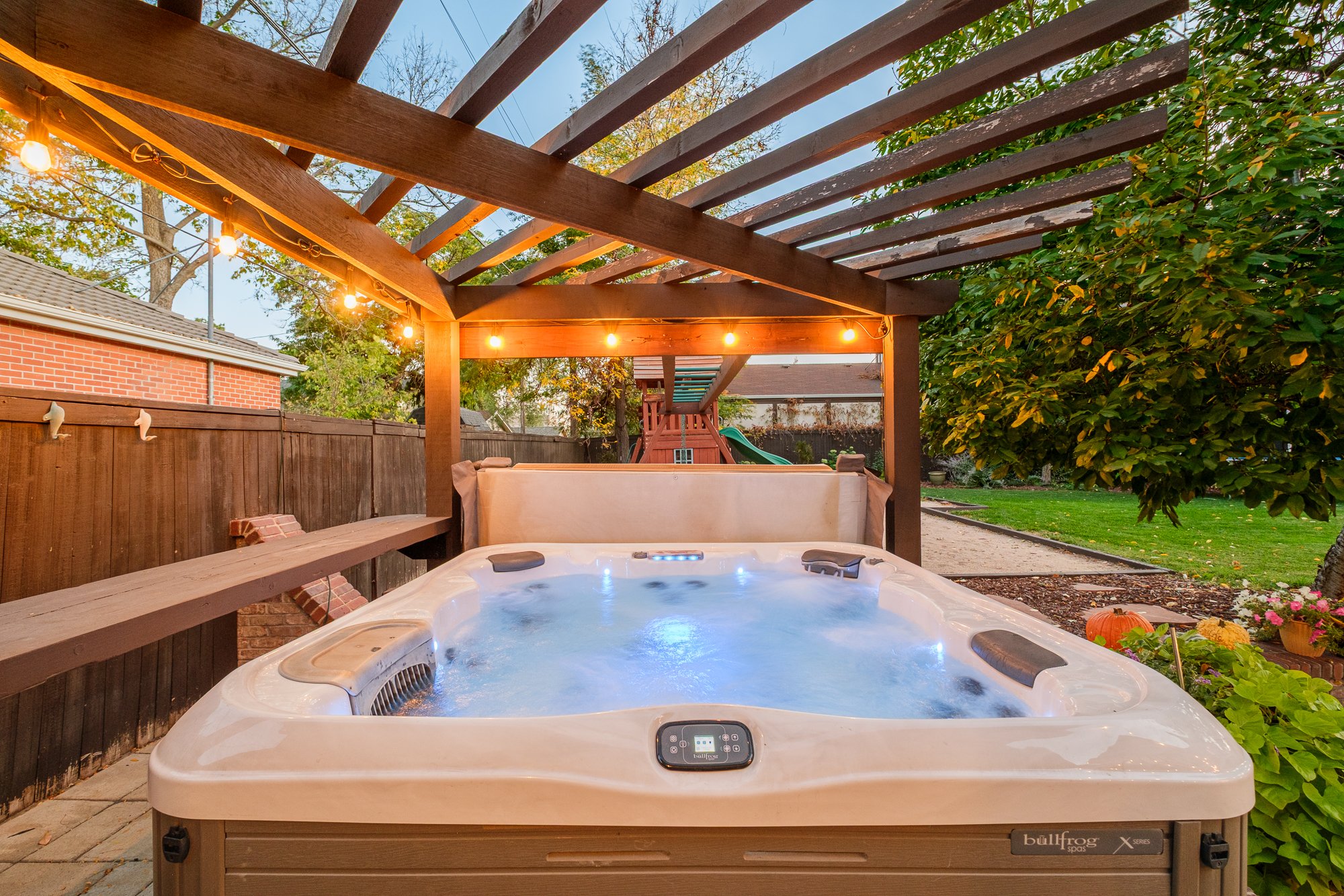 Outdoor hot tub beneath a wooden pergola with string lights in a landscaped backyard