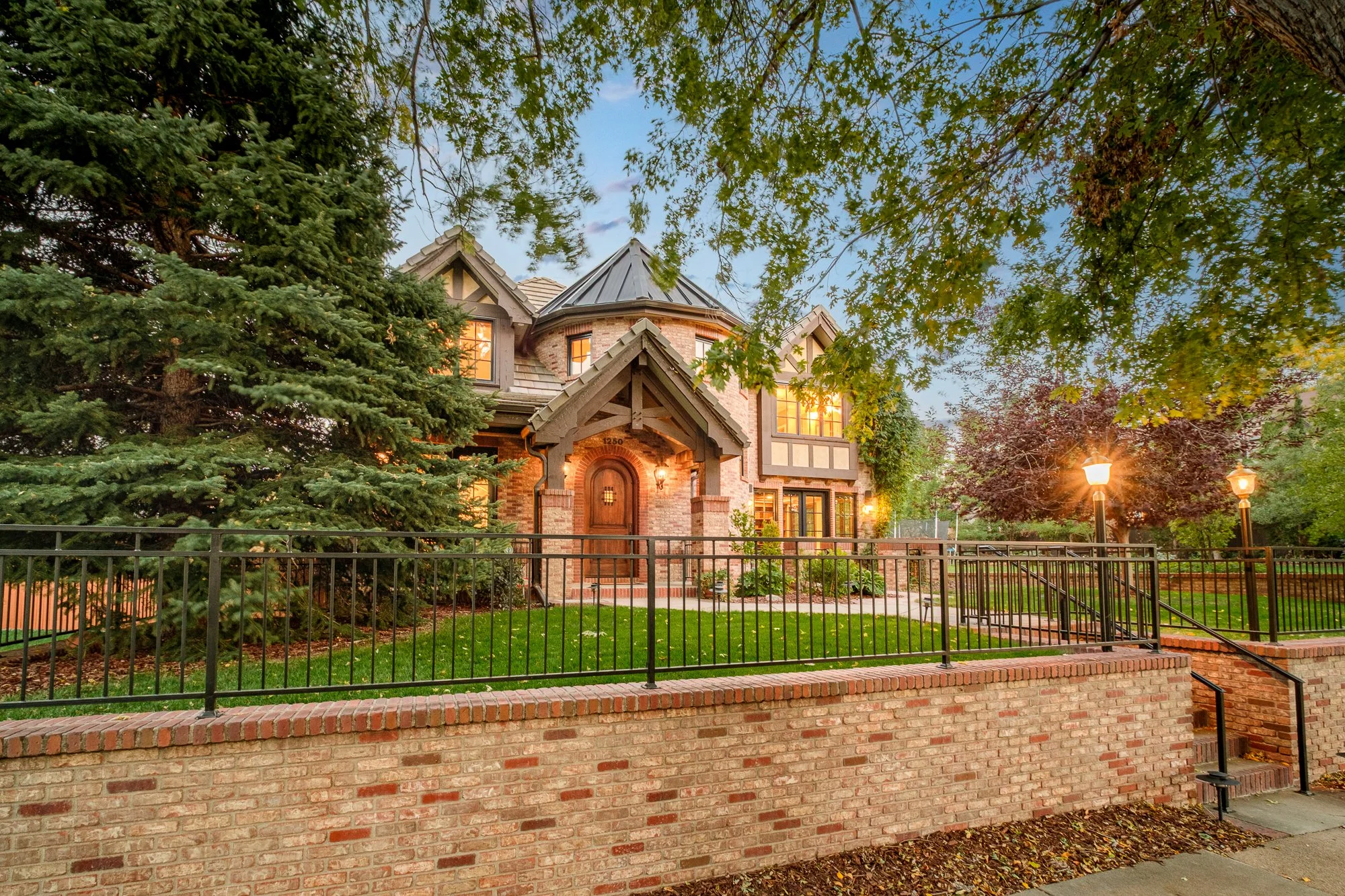 Large brick Tudor-style home with illuminated windows, landscaped yard, mature trees, brick retaining wall, and black metal fence at dusk