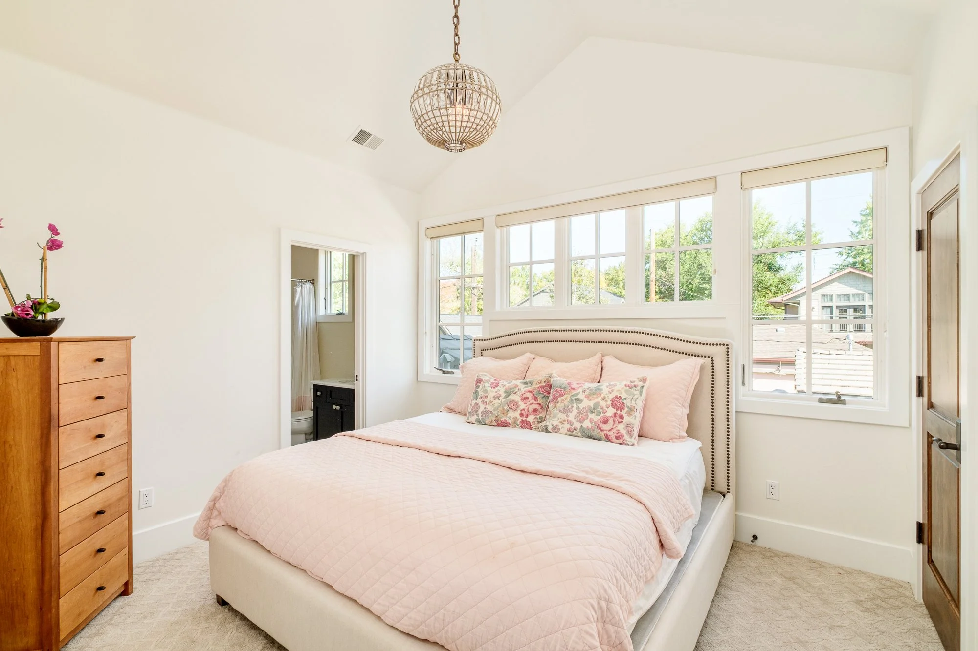 Bright bedroom with a pink quilted bed, floral pillows, large windows, wooden dresser, and hanging globe light fixture