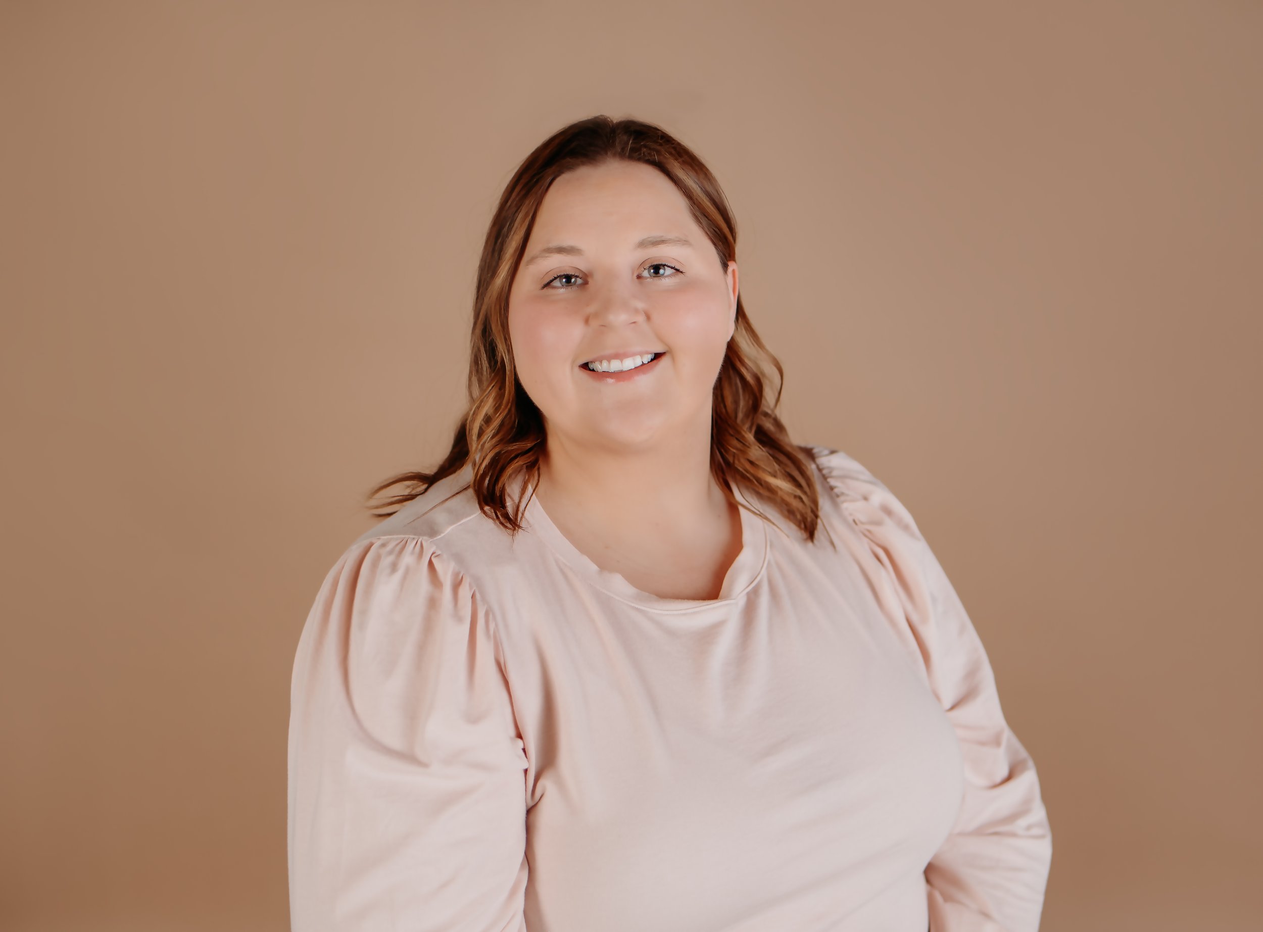 A young woman with wavy, shoulder-length reddish-brown hair, smiling, wearing a beige or light pink blouse with puffed sleeves, against a plain beige background.