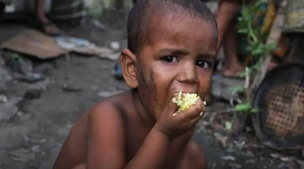 A young boy with dirt on his face and body, eating rice or grains, in a setting that appears to be in a poor area with debris and a woven basket in the background.