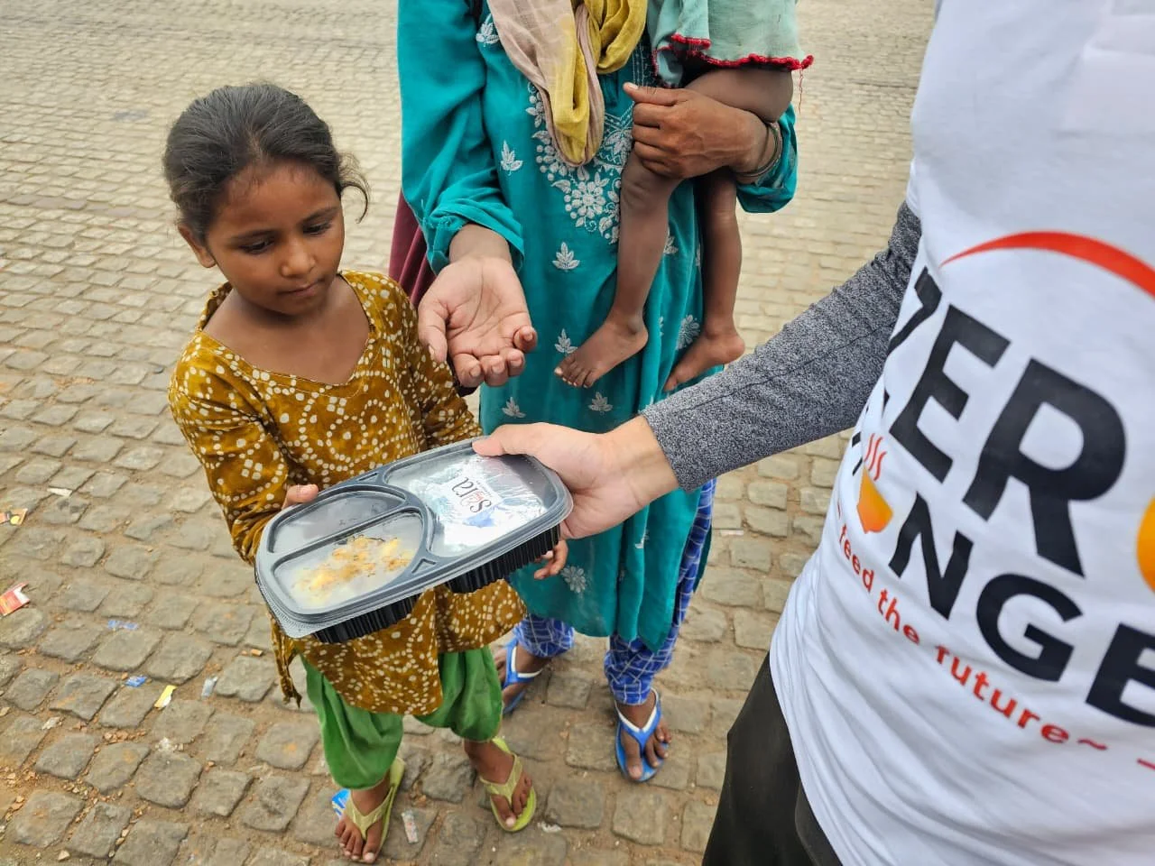 A young girl in a yellow patterned dress receiving a packed meal from a volunteer, with a woman holding a child in the background.