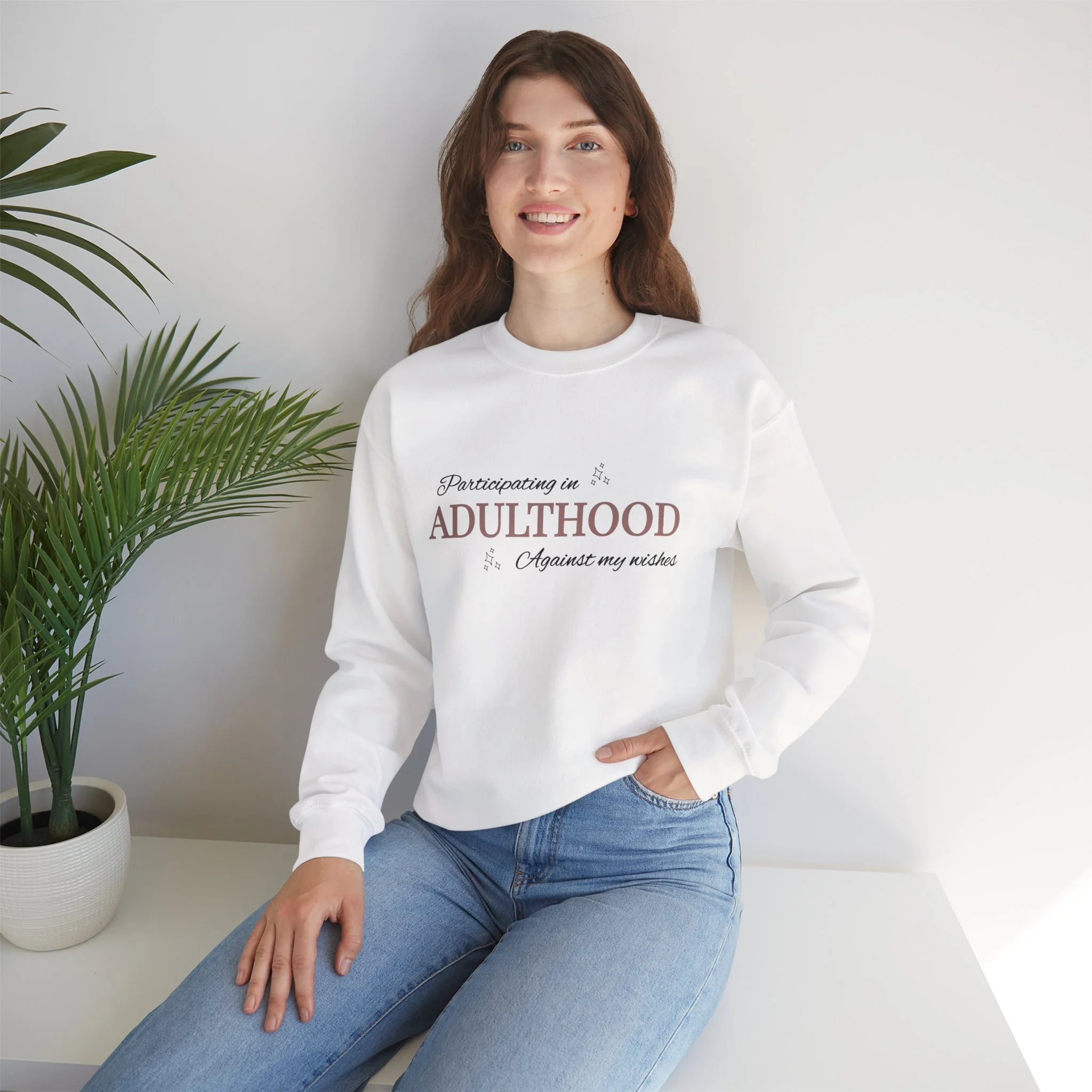 Young woman with curly brown hair, wearing a white sweatshirt with printed text, sitting on a white bench next to a potted plant with large green leaves, smiling at the camera.