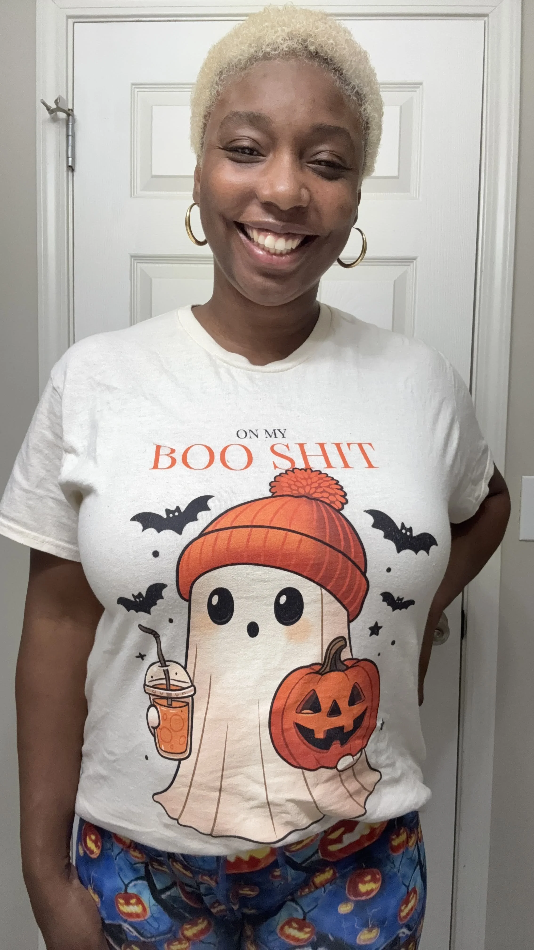 A woman with short, curly blonde hair and hoop earrings, smiling and wearing a white Halloween-themed T-shirt with a cartoon ghost holding a pumpkin and a drink, and pajama pants with a pumpkin pattern, standing in front of a door.