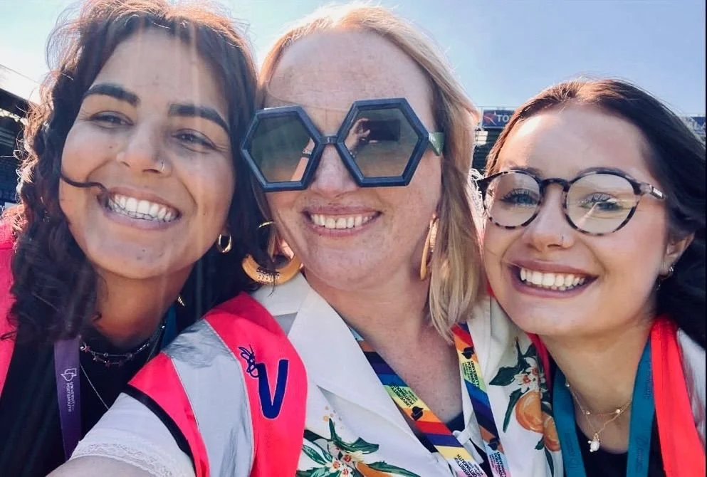 Three women smiling and taking a selfie outdoors during daytime, wearing colorful outfits and glasses, with a clear blue sky in the background.
