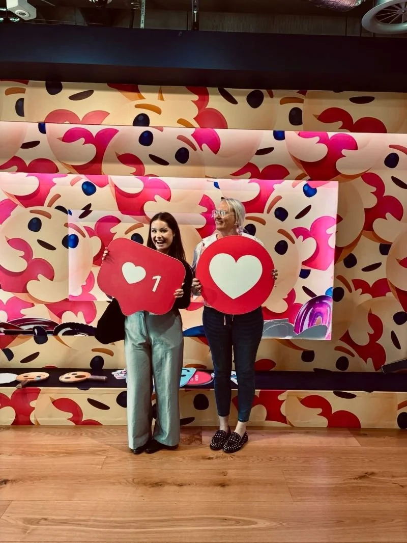 Two women standing in front of a wall decorated with pink, red, and beige smiley face pattern, holding large social media reaction icons; one with a heart symbol and the number 1, the other with a heart symbol.