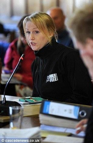 A young woman with blonde hair speaking into a microphone during a meeting, wearing a black jacket with a logo, with several people around her.