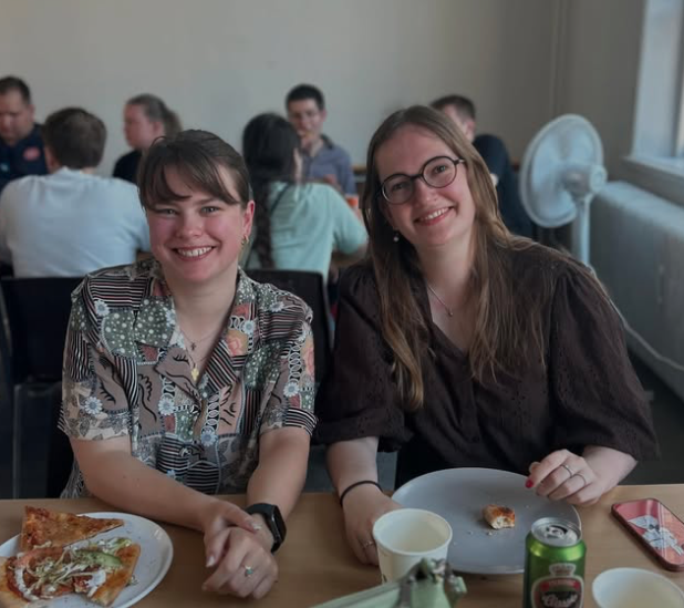Two young women sitting at a table in a crowded room, smiling at the camera. There are pizza slices, a can of soda, a paper cup, and a smartphone on the table.