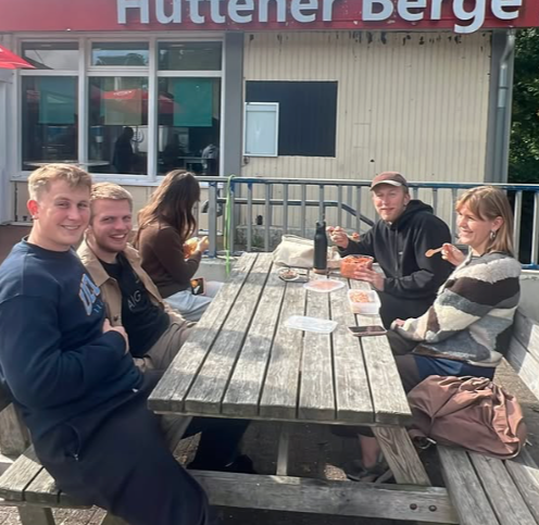 Group of five young adults sitting outdoors at a wooden picnic table, eating food and smiling, with a building and sign in the background.