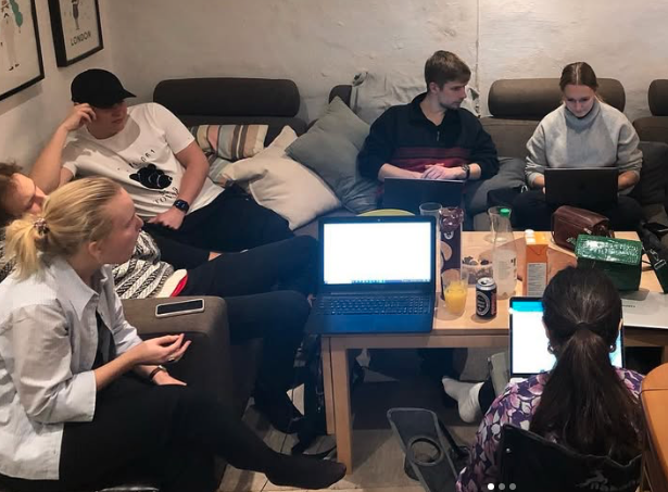 Group of six young people sitting around a table with laptops and drinks in a cozy room with cushions and framed artwork on the wall.
