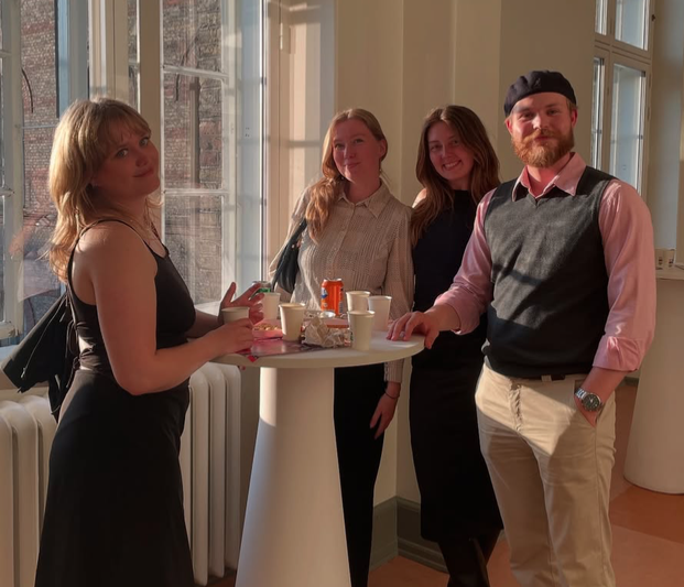 Four young adults at a standing table with drinks and snacks in a sunlit room with large windows.