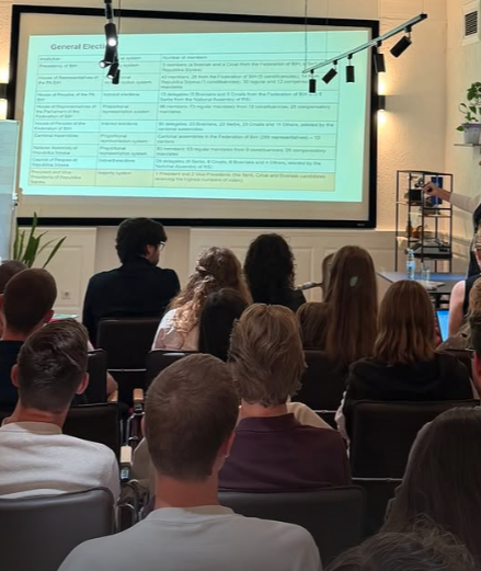 A group of people watching a presentation in a conference room with a large projected slide on the wall, and a person on the side taking photos.