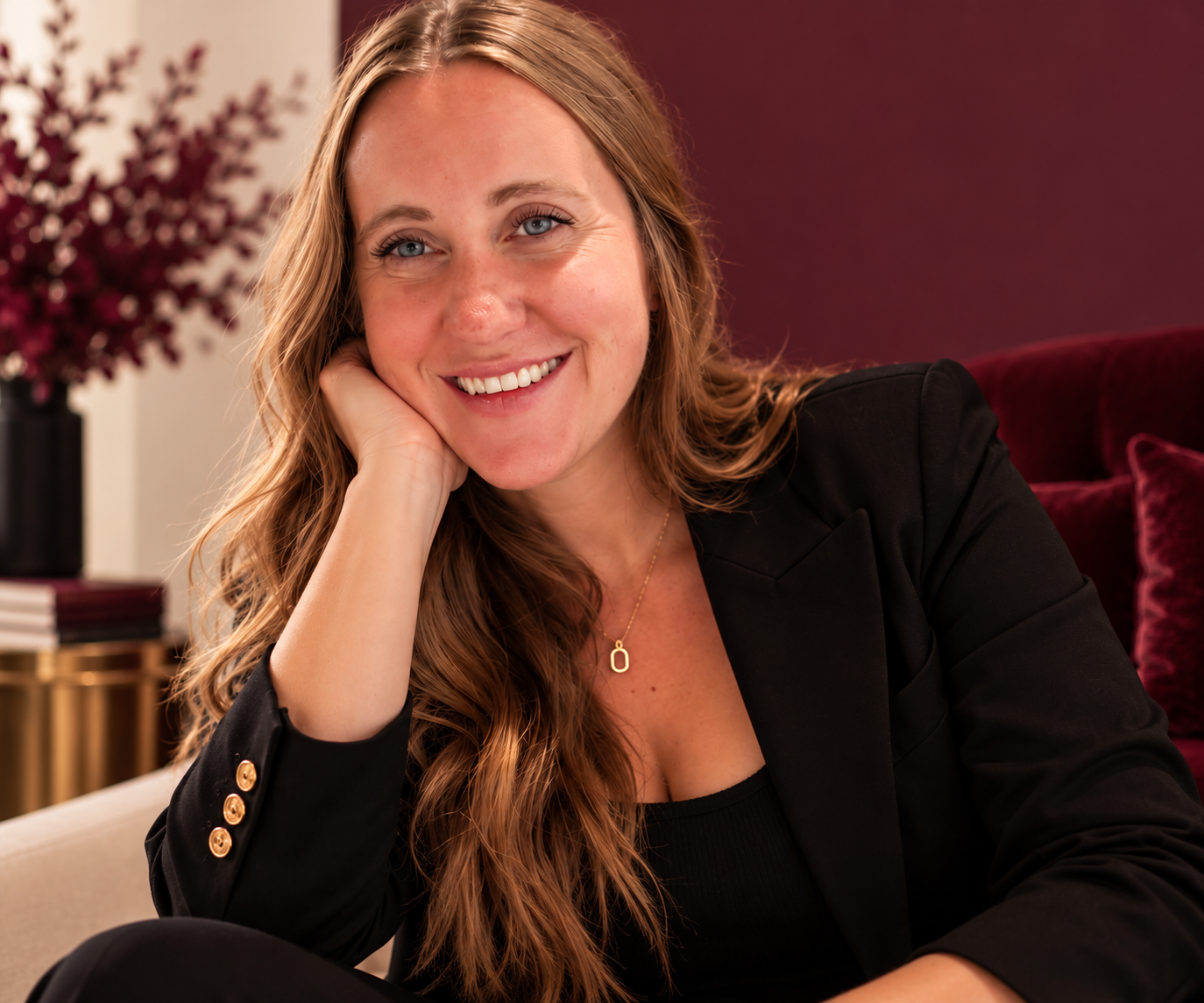 Smiling woman with long blonde hair wearing a black blazer and gold necklace, seated on a burgundy sofa