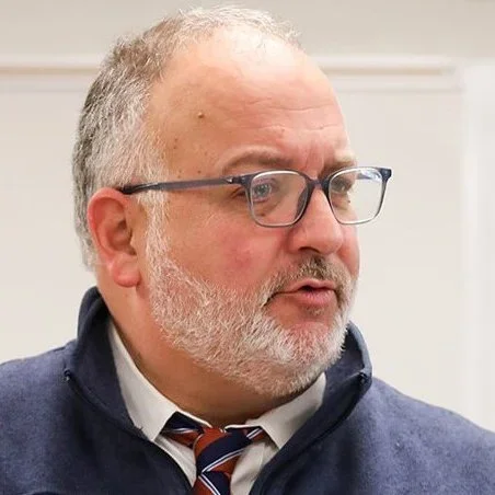 Close-up portrait of a gray-haired, bearded man wearing glasses, a collared shirt, striped tie, and navy jacket.