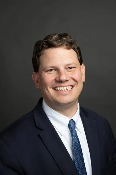 Professional headshot of a smiling man in a navy suit and blue tie against a dark gray background