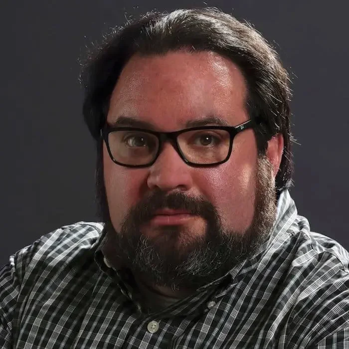 Portrait of a bearded man wearing black glasses and a plaid shirt against a dark background