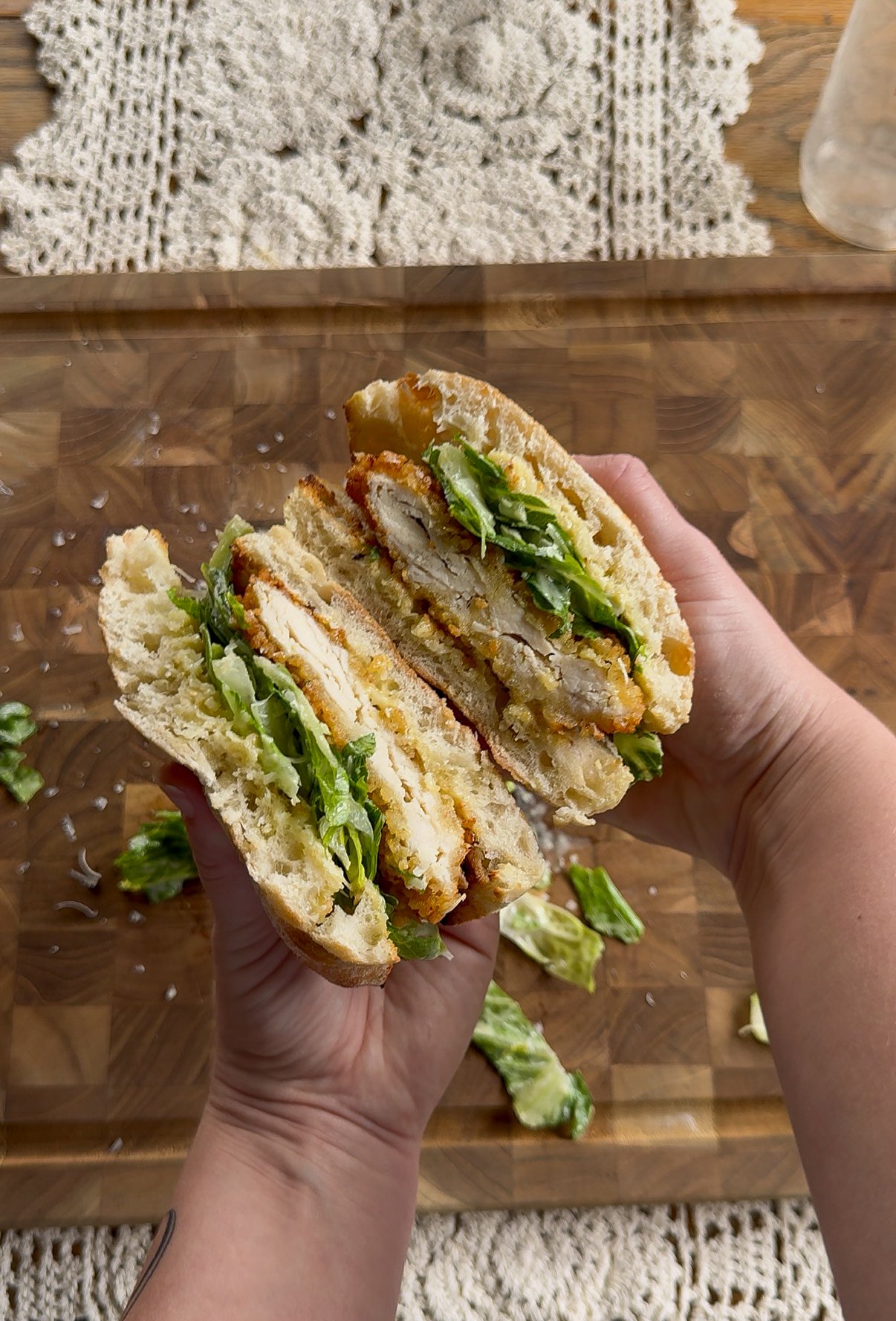 Hand holding a crispy chicken sandwich with lettuce on a wooden cutting board