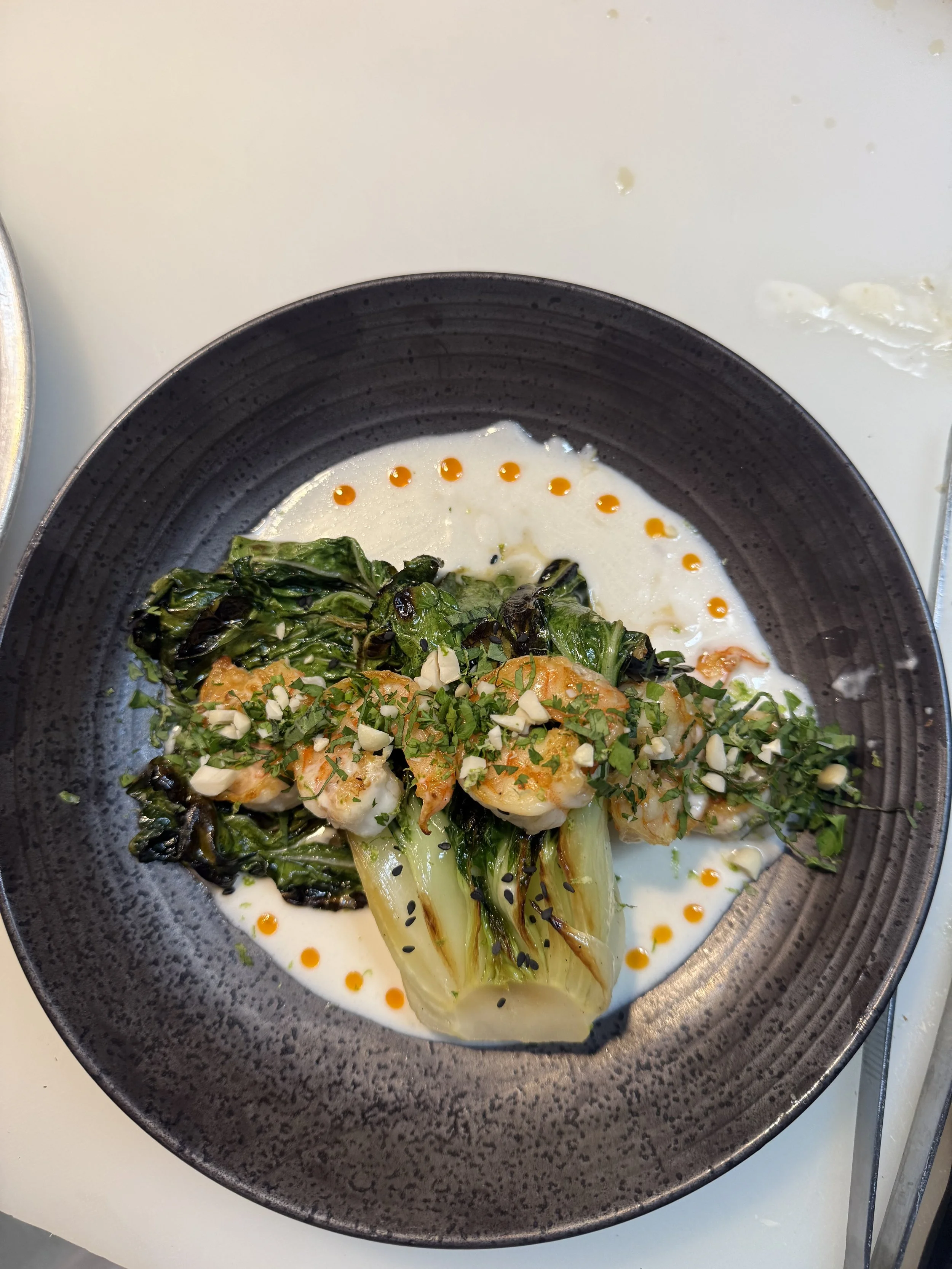 Close-up of grilled shrimp and bok choy with roasted greens, tomatoes, herbs, almonds, black sesame seeds, and orange sauce dots on a dark plate.