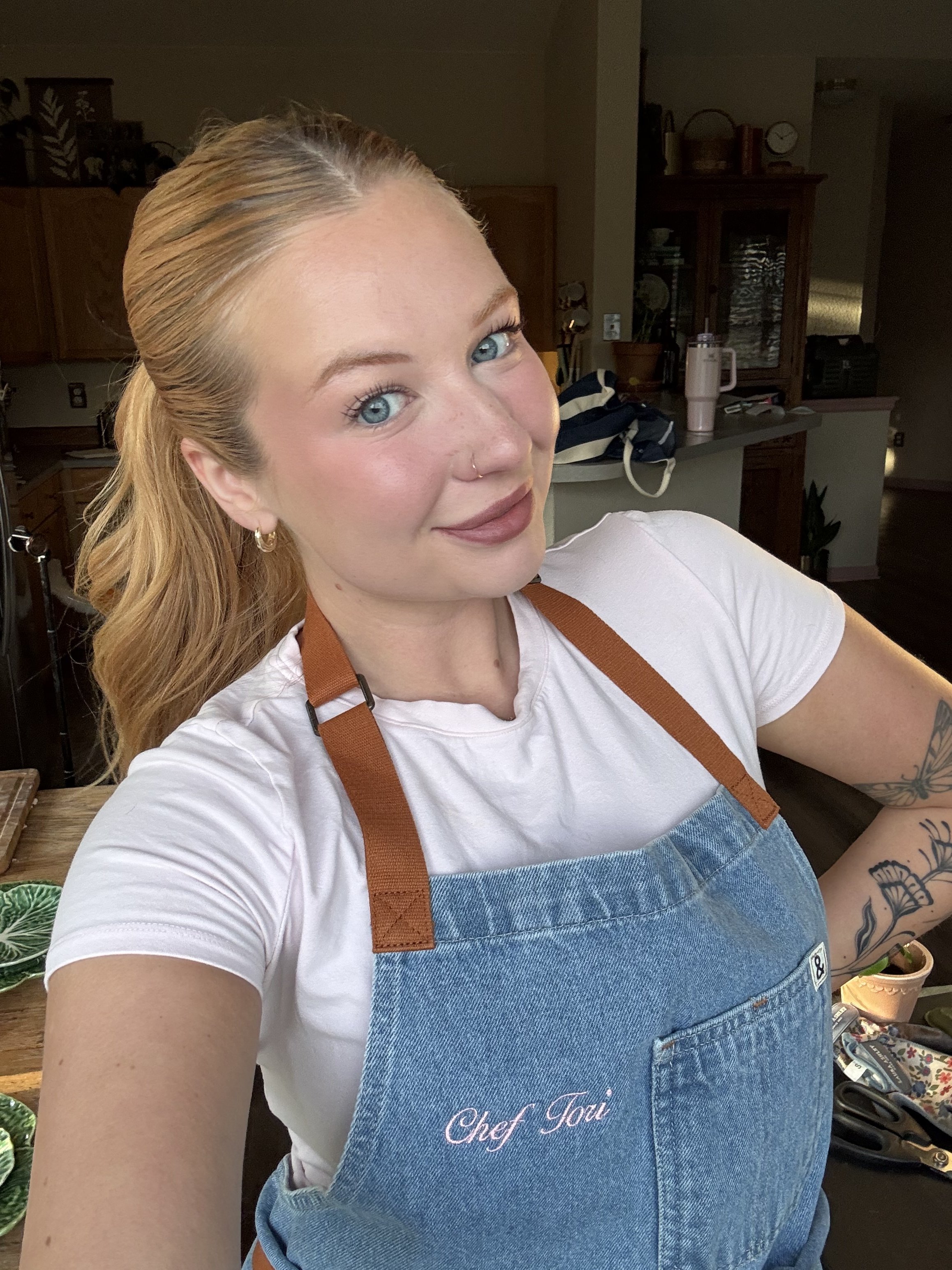 Close-up selfie of a smiling blonde woman wearing a white T-shirt and blue denim apron embroidered with “Chef Toni” in a home kitchen.