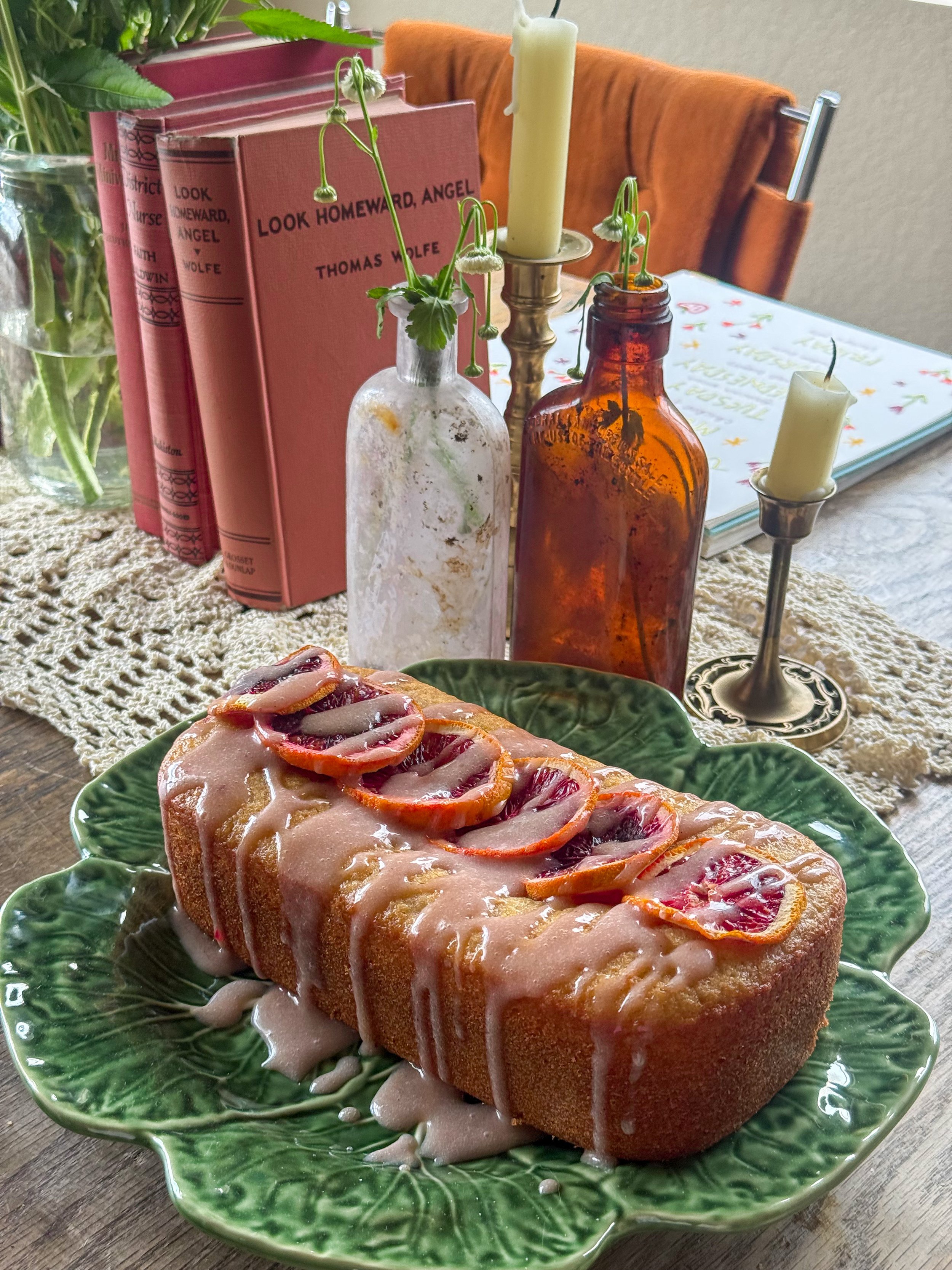 Glazed Bundt cake topped with blood orange slices on a green leaf-shaped serving plate, with candles, flowers, and books in the background.