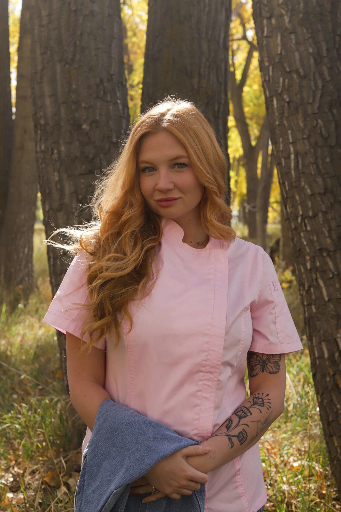 Woman with long red hair wearing pink chefs coat and holding a folded blue garment in a sunlit forest