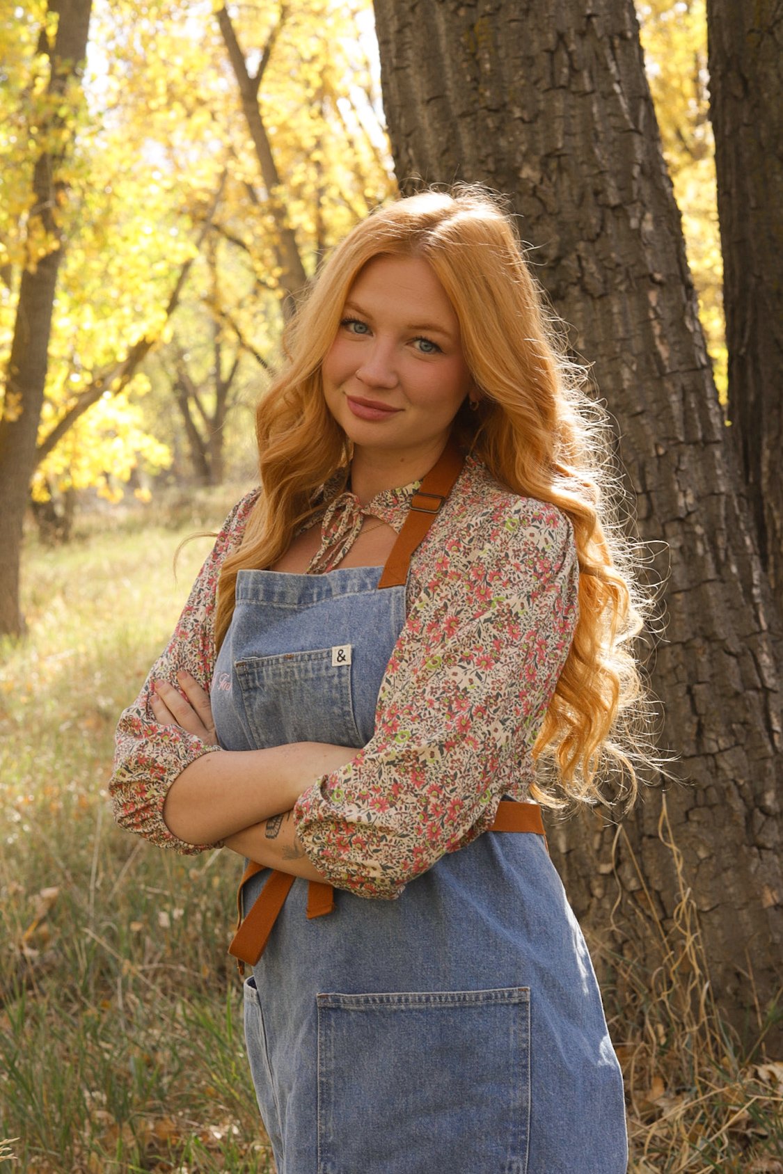 Woman with long red hair wearing a floral blouse and denim apron standing in a sunlit autumn forest