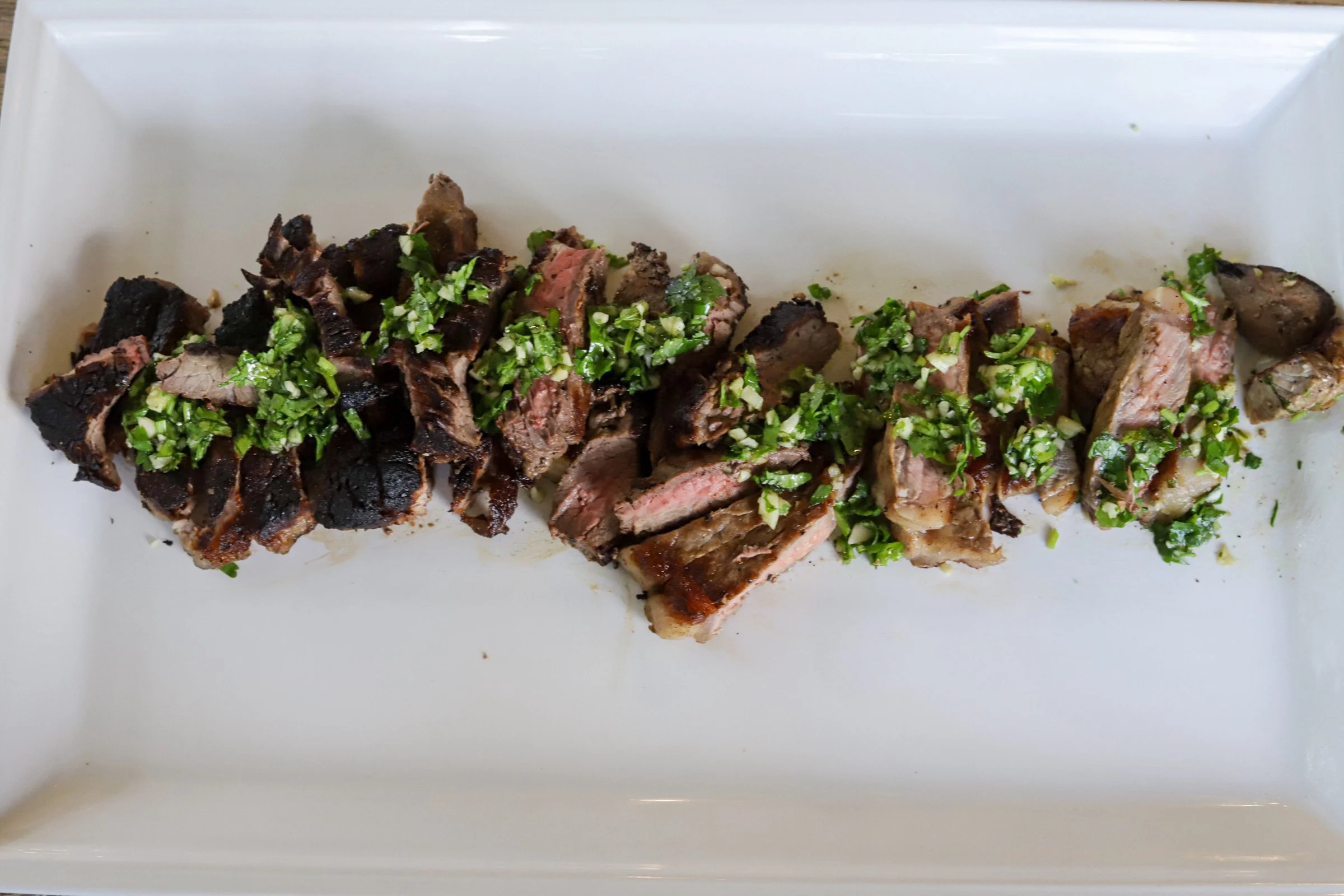 Sliced grilled steak topped with chopped parsley and garlic on a white serving platter
